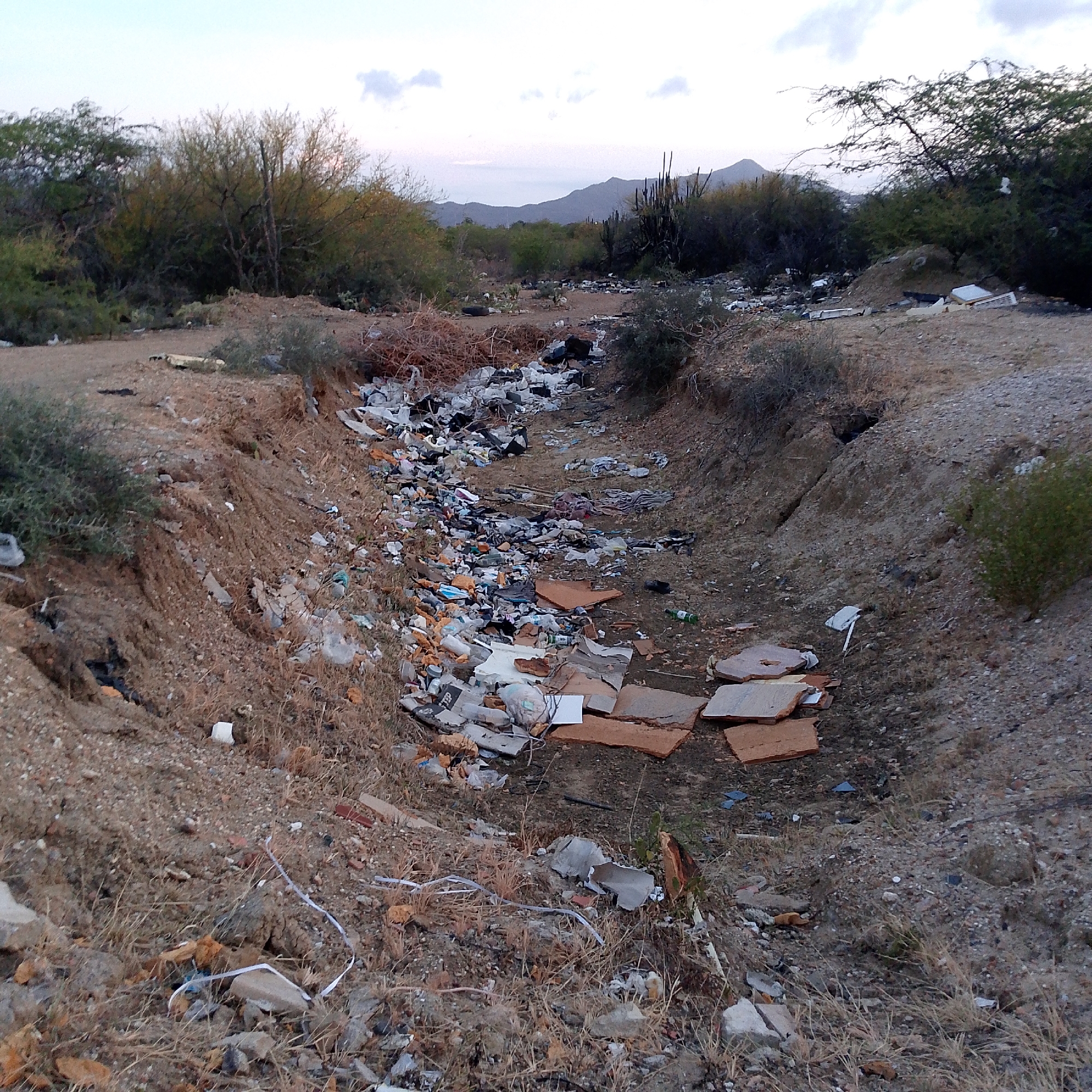A ditch filled with trash runs through a dry, desert-like landscape. The trash consists of plastic bottles, styrofoam, pieces of tile, and other debris. Bushes and trees are present on both sides of the ditch. In the background, there are mountains. The location appears to be a rural area, possibly on the outskirts of Altagracia, Venezuela. There are no people visible in the image.