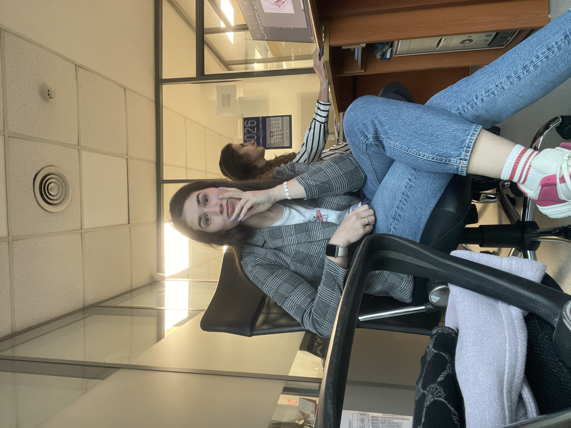The image depicts two young women indoors, likely in an office setting in Sofia, Bulgaria. The location is evidenced by the Cyrillic calendar on the wall displaying the year 2026, typical of Bulgaria. The woman in the foreground is seated in a black office chair wearing blue jeans, a grey checkered blazer, a white shirt, and pink-and-white sneakers with red stripes on the socks. She is making a gesture with her hand. The second woman, sitting behind and to the left, is wearing a striped long-sleeved shirt and appears to be looking at a computer screen. The office environment includes a suspended ceiling, glass partitions, office furniture, and equipment.