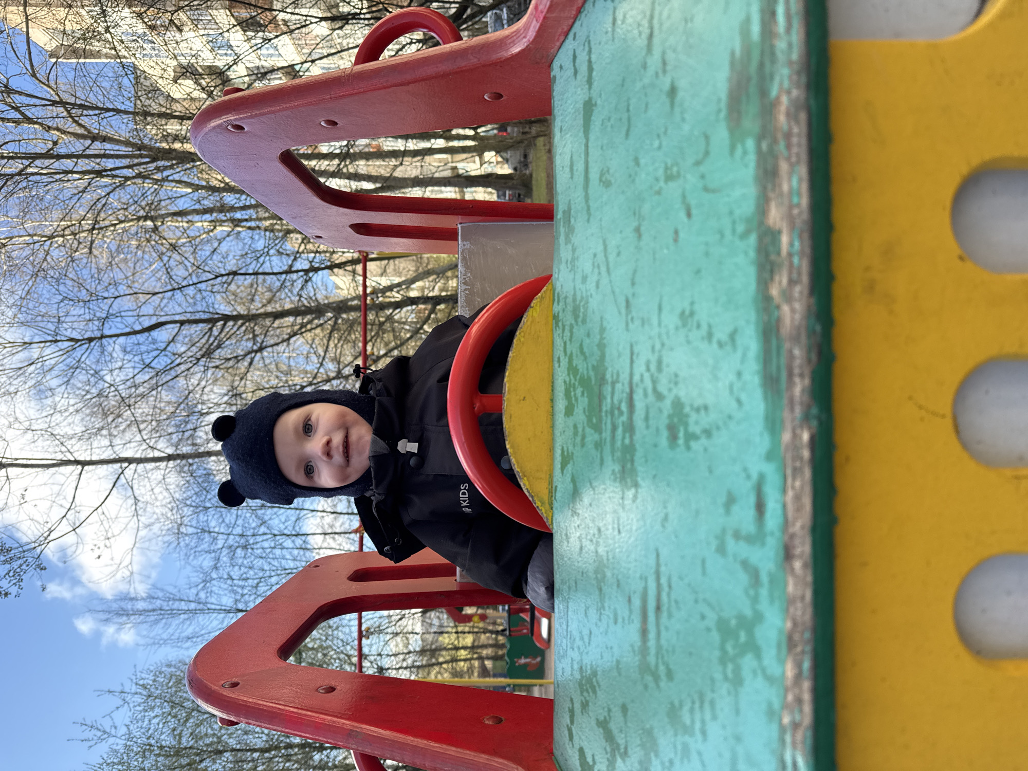 A young child wearing a dark blue hat with pom-poms and a black jacket is centered in the frame, looking directly at the camera with a slight smile. The child is holding a red steering wheel, part of a playground structure, and appears to be playing on it.
The playground equipment includes red, turquoise, and yellow painted wood, showing signs of wear and tear. The background is blurred but shows trees without leaves and a blue sky with light clouds, suggesting a late autumn or early spring day. Buildings are visible through the trees, indicating an urban environment.
The location is Saint Petersburg, Russia.