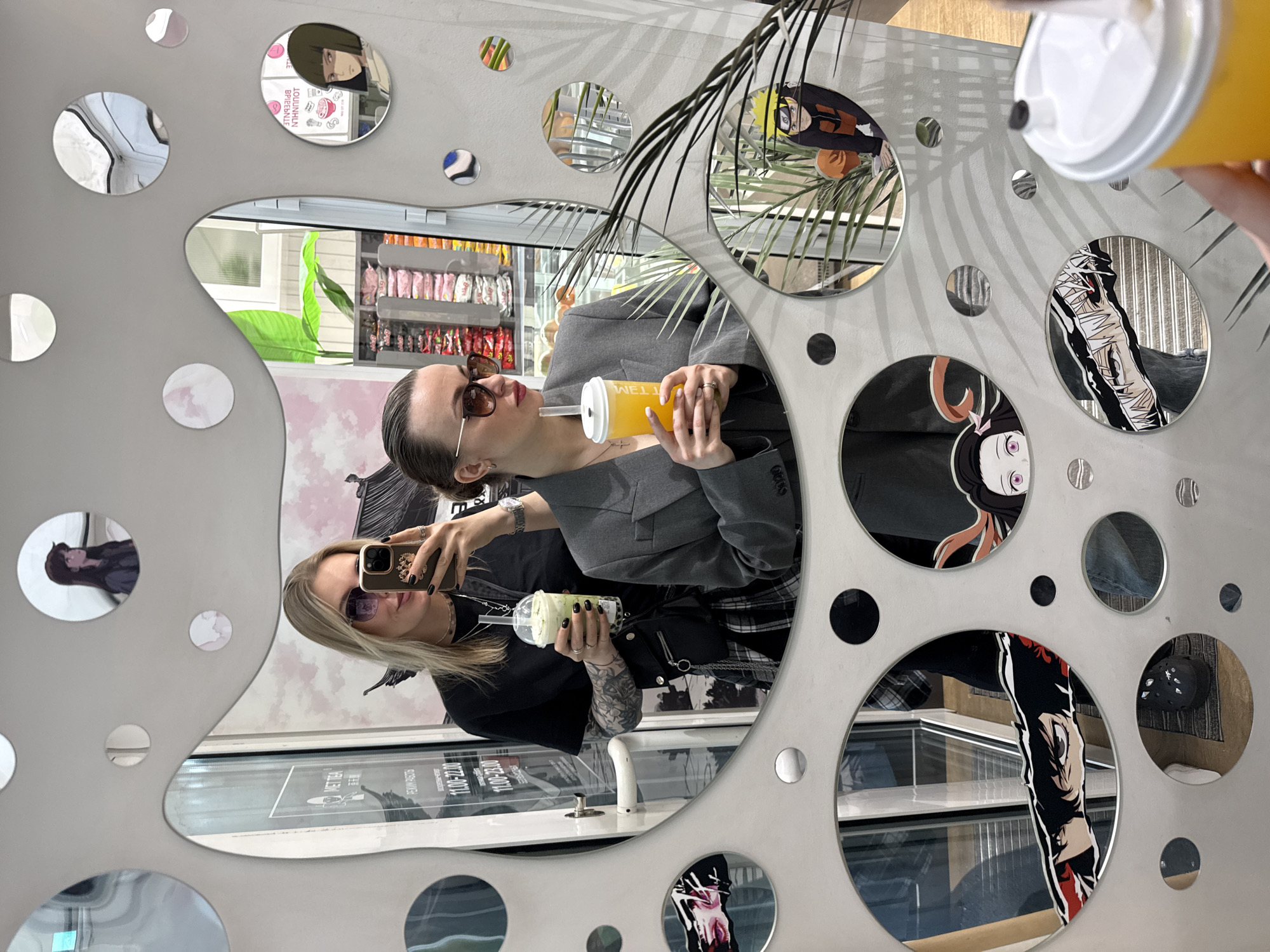 The image shows two women reflected in a decorative mirror in a public space, likely a cafe or shop. The mirror has multiple circular cutouts, some filled with anime character stickers. The woman closer to the camera is holding a phone and appears to be taking a selfie. She has blonde hair and wears sunglasses and a black t-shirt. The other woman is wearing a grey blazer and holding a beverage. Behind them, shelves of merchandise are visible. The mirror also reflects part of the exterior environment. Based on the mirror decor and architecture, it is likely in Krasnoyarsk, Russia.