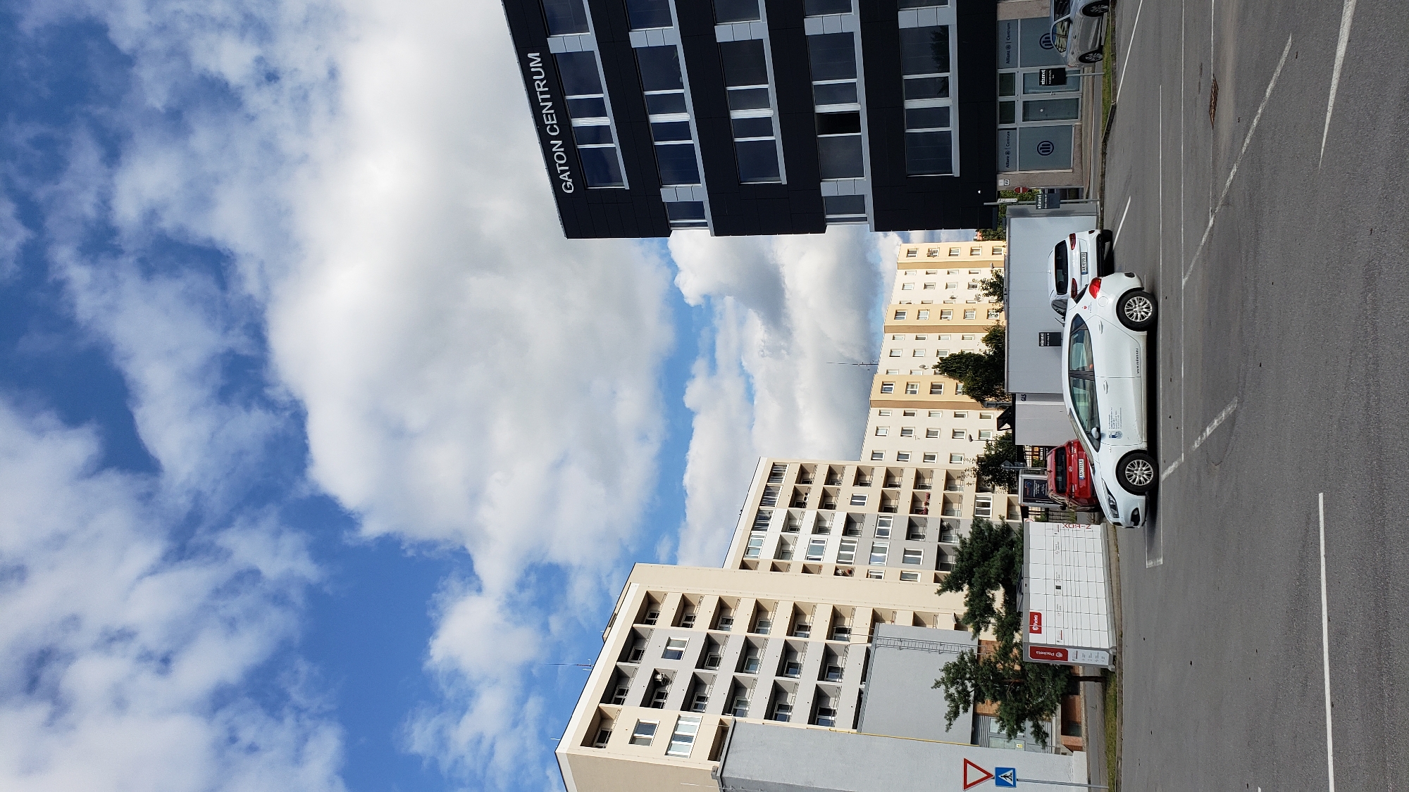The image depicts a daytime exterior scene in Prešov, Slovakia, focusing on a parking lot adjacent to several buildings. In the foreground, a white hatchback car is parked, and a red car is visible behind it. To the left, a tall, light-colored apartment building dominates the frame, featuring numerous balconies. Further to the right, a modern, dark-colored office building with large windows displays the signage "GATON CENTRUM". Beyond the apartment building, another row of light-colored buildings, appearing to be residential, stretches into the background. The sky is partly cloudy, with patches of blue sky visible between white clouds. The ground is paved asphalt, marked with parking spaces. A white parcel locker station is situated near the apartment building. The overall environment suggests an urban setting with a mix of residential and commercial structures.