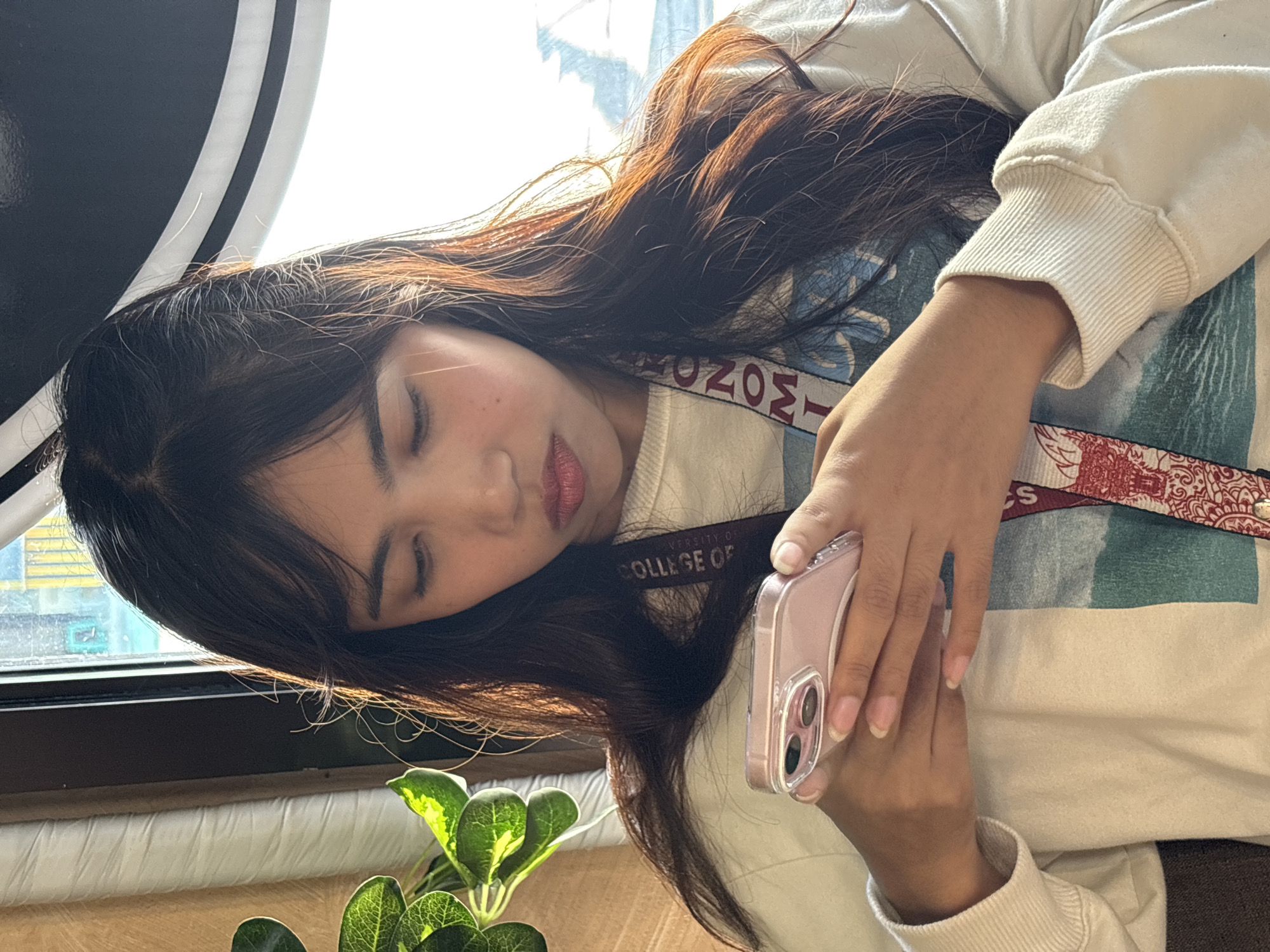 A young woman with long, dark hair is shown from the chest up, looking down at her pink smartphone. She is wearing a light-colored sweatshirt with a design featuring a blue and green landscape. A lanyard with text, possibly a university name like "COLLEGE OF ECONOMICS," is visible around her neck and draped across her chest. She is seated indoors, with a window to her right that allows sunlight to illuminate her hair and face. To her left, a portion of a green plant with large leaves is visible. The background suggests an indoor setting, possibly a cafe or a room with a window overlooking an outdoor scene. The image does not provide any specific landmarks or context that would allow for the identification of its location within Davao City, Philippines.
