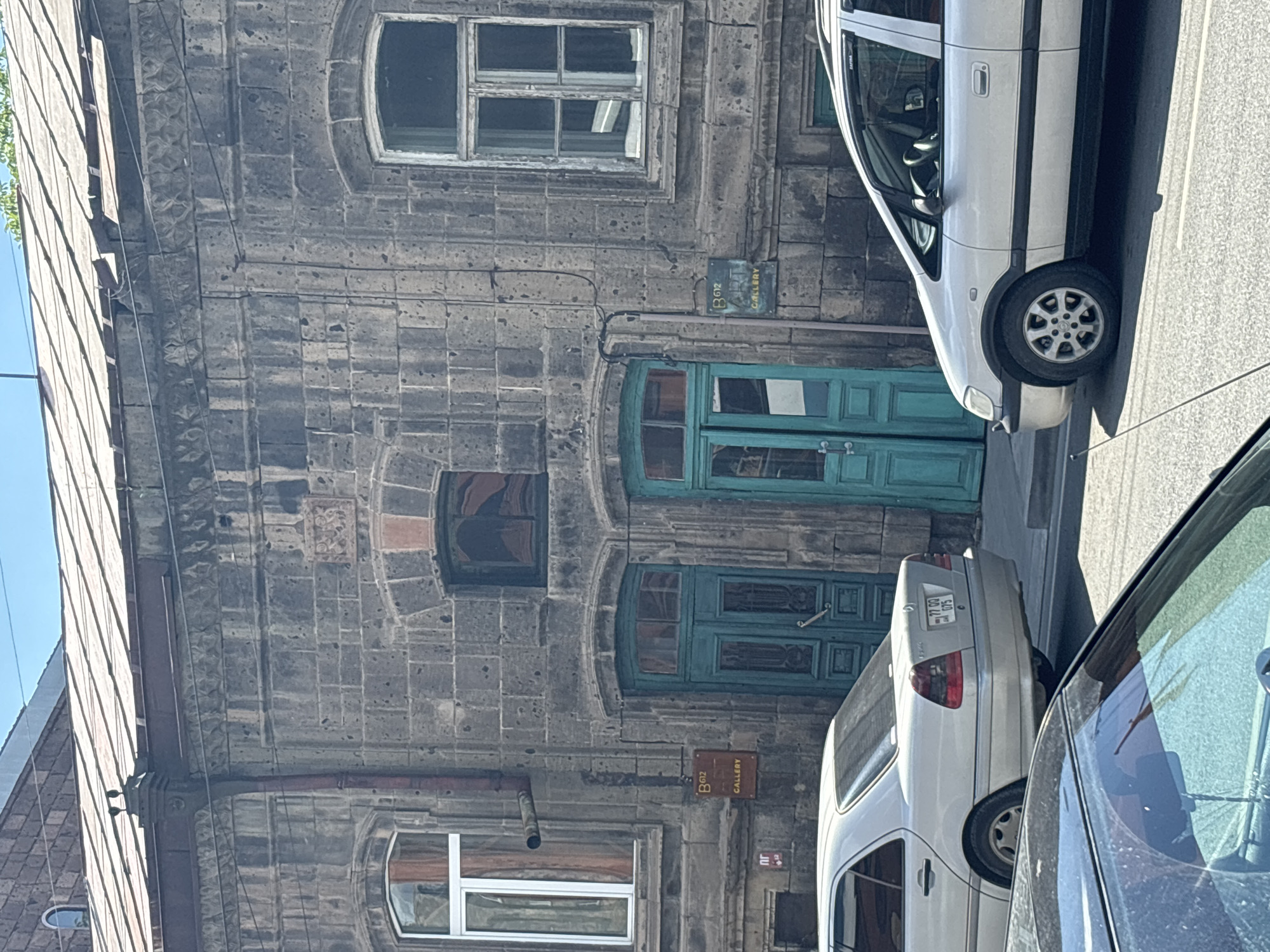 The image shows the facade of a building constructed with dark gray stone blocks. The building has several windows, some with arched tops, and two prominent teal-colored doors with ornate detailing. A small, brown sign with "B012 GALLERY" is mounted on the wall between the two doors. Electrical wires are visible running across the facade.
In the foreground and right side of the image, parts of two cars are visible. A silver sedan is parked next to the building, and a darker car is partially visible in the far right. The silver sedan has a license plate with characters that appear to be Cyrillic.
The scene appears to be outdoors on a sunny day, indicated by the strong shadows cast by the cars and the bright sunlight reflecting off the building and vehicles. The architecture suggests an urban setting, possibly in Eastern Europe or a region with similar architectural styles. No people are visible in the image, and the overall mood is neutral, focusing on the architectural details and the urban environment. The time of day is likely midday due to the angle of the shadows.