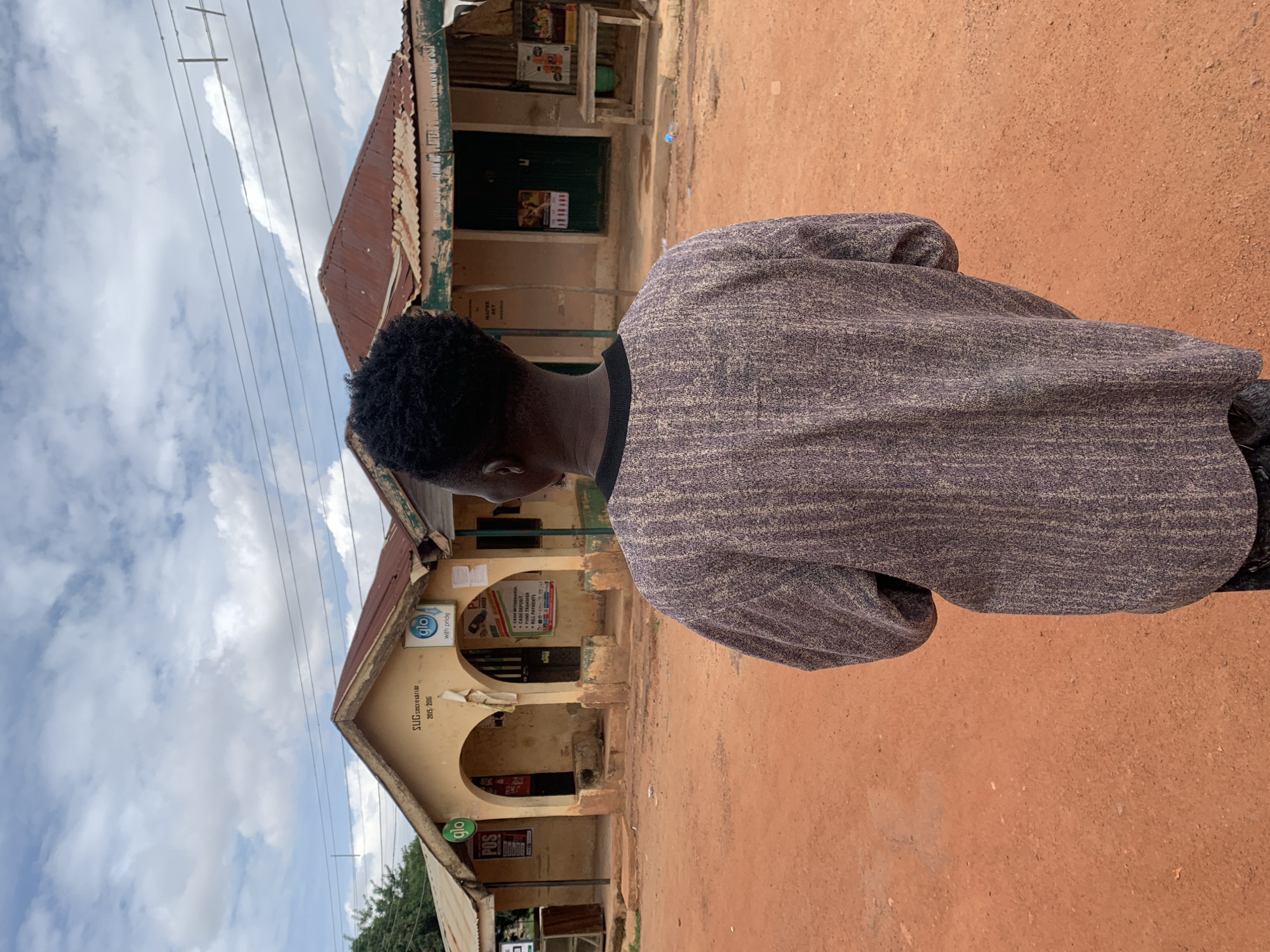 The image captures a person standing on a reddish-brown dirt ground, viewed from behind, facing two low-rise buildings under a partly cloudy sky. The scene is located in Jos, Nigeria.
The foreground is dominated by the back of an individual wearing a textured, long-sleeved garment with vertical patterns in shades of purple-grey and light brown. A dark collar is visible at the neck. The person has short, dark, curly hair. The lower portion of dark trousers or a skirt is barely visible at the bottom edge.
The midground features two adjacent, single-story buildings constructed of light brown/beige plaster. Both structures have corrugated metal roofs, which appear rusty and reddish-brown. The central building features two arched entrances. Above the left archway, a sign reads "SUG Secretariat" with "2015, 2016" printed below. Multiple "glo" telecommunications company signs are affixed to the buildings; one on the left facade displays "glo POS," while another on the central building states "glo with pride." Posters and flyers are attached to the building walls, including a white poster on the central building listing services such as "CASH WITHDRAWAL," "DEPOSIT MONEY," "FUND TRANSFER," and "AIRTIME PAYMENTS." A dark green door is visible on the rightmost building, beside which a simple wooden table or bench stands. The ground is composed of reddish-brown soil, typical of laterite, with small scattered debris.
The background consists of a sky with dispersed white and grey clouds. Several dark power lines or communication cables traverse the upper portion of the frame horizontally, with some vertical supports visible. Green foliage from distant trees or bushes can be observed on the far left edge of the image.