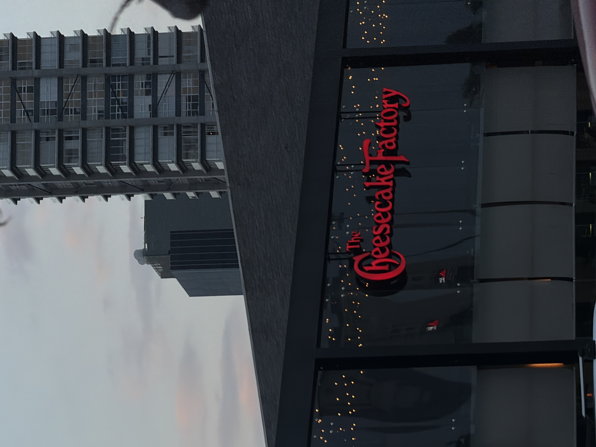 The image, taken in Puebla, Mexico, presents an urban landscape dominated by two distinct architectural structures under a soft, light gray sky, suggesting either an overcast day or the transition into evening.
On the right side of the frame, a prominent, dark-toned building houses "The Cheesecake Factory." Its large, red, stylized script sign, reading "The Cheesecake Factory," is vibrantly illuminated and clearly displayed on a wide glass facade. Behind the glass, numerous small, warm-toned lights twinkle, creating an inviting and festive atmosphere, characteristic of the restaurant's interior or storefront decoration. The building's exterior appears to be constructed from dark stone or a similarly textured material.
To the left, a tall, modern building stands with a striking architectural design. It features multiple stories characterized by a repetitive pattern of rectangular windows and dark, horizontal elements that create a grid-like effect, possibly balconies or sun shades. Below this larger structure, a smaller, dark building with a flat roof is partially visible.
The scene focuses purely on the architecture; no specific human activity or people are visible. The artificial illumination of "The Cheesecake Factory" contrasts with the subdued natural light, reinforcing the impression that the photograph was taken during late afternoon or early evening. The overall mood is calm, highlighting the juxtaposition of a commercial establishment's warm glow against the backdrop of more austere urban structures.