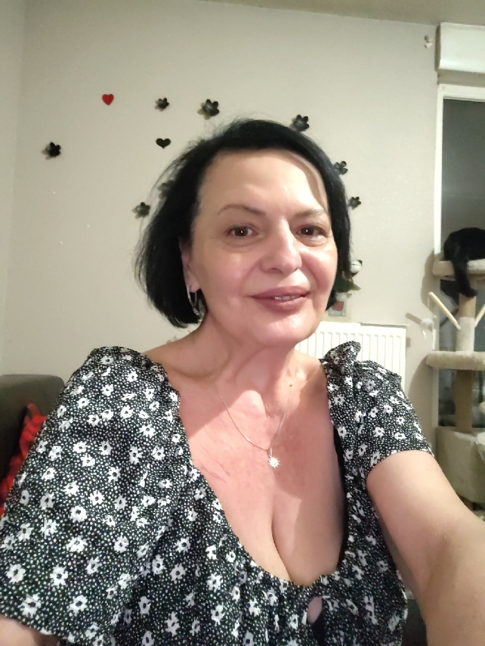 The scene depicts an adult female in an indoor residential setting, located in Ostwald, France. The female, appearing middle-aged, is positioned centrally, looking directly at the camera with a slight smile. She has short, dark hair, a light complexion, and is wearing a black dress with a white floral pattern, featuring a wide, open neckline and short, ruffled sleeves. A silver-toned necklace with a circular, radiating pendant hangs around her neck, and she wears small silver-toned hoop earrings.
The background wall is light-colored and decorated with multiple dark, flower-shaped appliques and two red heart-shaped appliques. To the subject's left, a portion of a dark-colored sofa with a red and black plaid cushion is visible. To her right, a white panel radiator is present. Further to the right, a multi-tiered, light-colored cat tree is visible, upon which a black cat is resting, facing away from the camera. A window or glass door with a white frame and a partially lowered light-colored blind is discernible in the far right background.