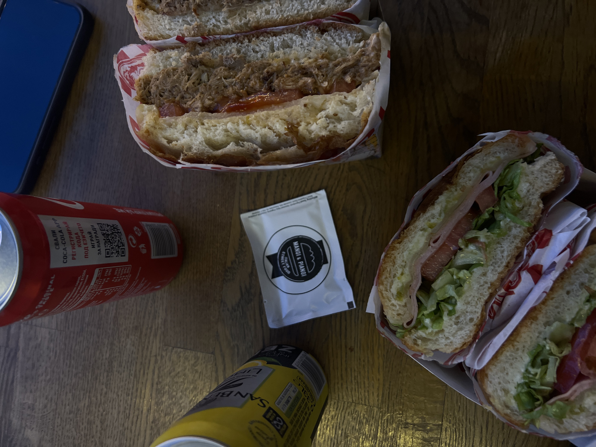 The image shows a wooden table surface with several food and drink items. There are two halves of what appear to be pulled pork sandwiches in red and white paper wrapping and two halves of ham and cheese sandwiches with lettuce and tomato in similar wrapping. A white sachet with a logo "Mangi & Piangi ITALIAN STREET FOOD" is visible between the sandwiches. A can of Coca-Cola is placed on the left of the sachet. A yellow can of what appears to be San Pellegrino Limonata is at the bottom of the sachet. A blue smartphone is partially visible on the top left corner of the image. The items on the table suggest a meal is being consumed. The logo on the sachet indicates the location may be at or associated with an Italian street food vendor called "Mangi & Piangi", potentially in Sofia, Bulgaria as suggested by the Coca Cola text, although this is not confirmed in the image itself.