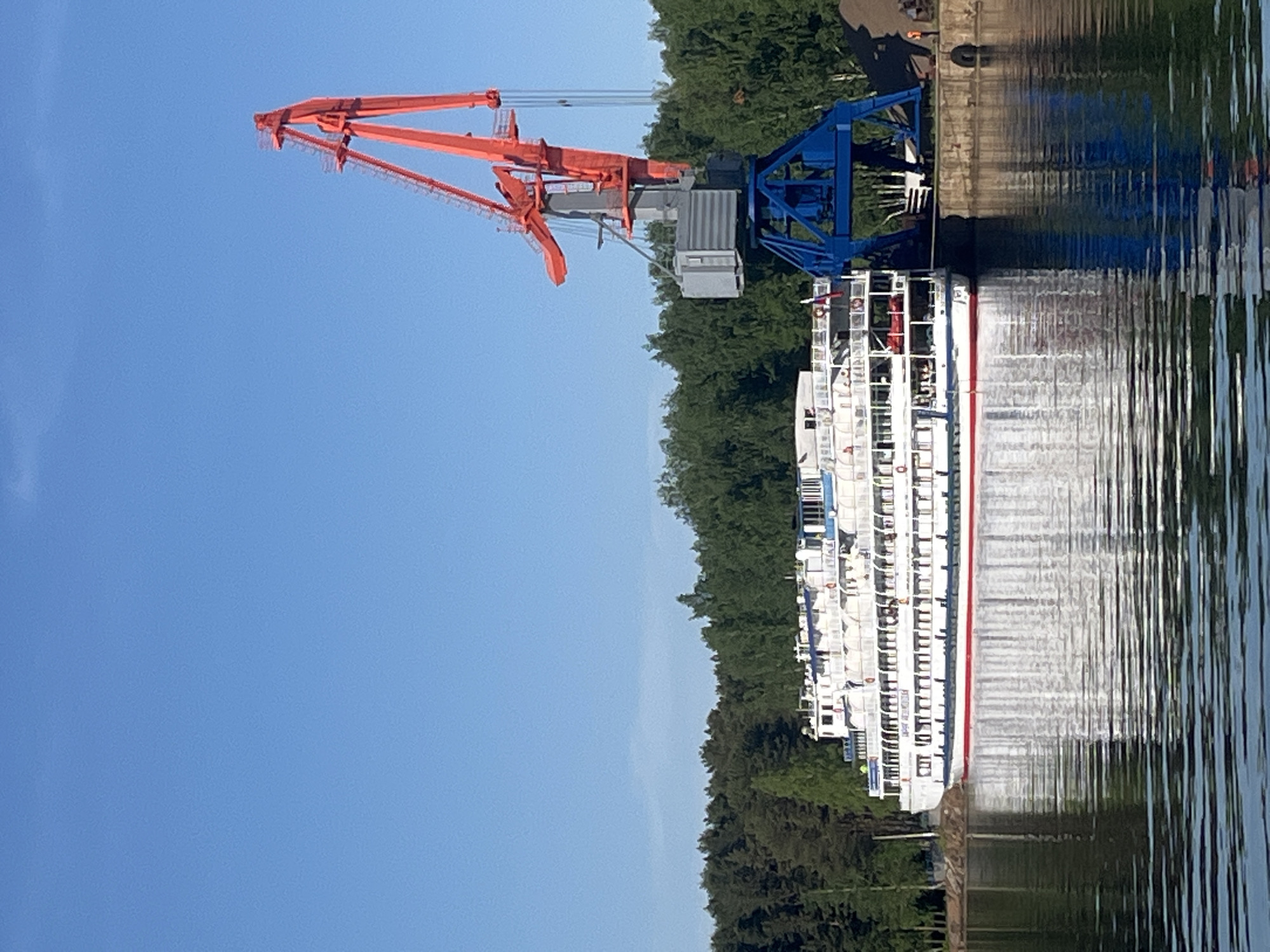 The image depicts a river scene in Chaikovsky, Russia. A large, white river cruise ship with a red stripe is docked along a concrete pier. The ship's reflection is visible in the water. A large crane with an orange boom and blue supports is positioned next to the ship, likely used for loading or unloading. A dense line of green trees stretches along the riverbank behind the ship and crane. The sky is clear and blue.