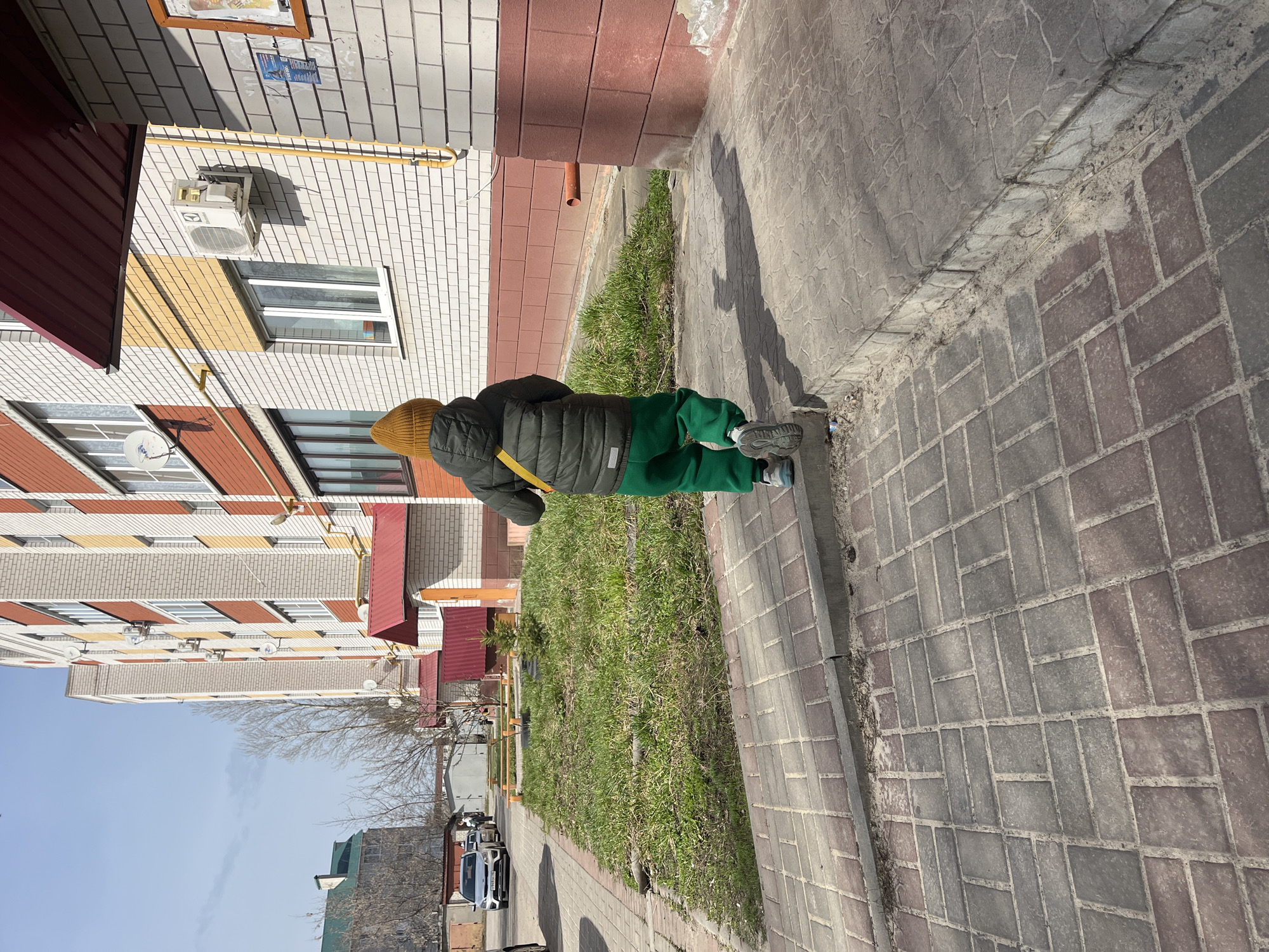 A child wearing a yellow knit hat, a black puffer jacket with a yellow strap, and green pants stands on a brick sidewalk next to a grassy area. Adjacent to the grass is a multi-story building with brick and siding. The building has air conditioning units and windows. Beyond the grass, a street with parked cars is visible. The sky is blue. The photo was taken in Usman, Russia.