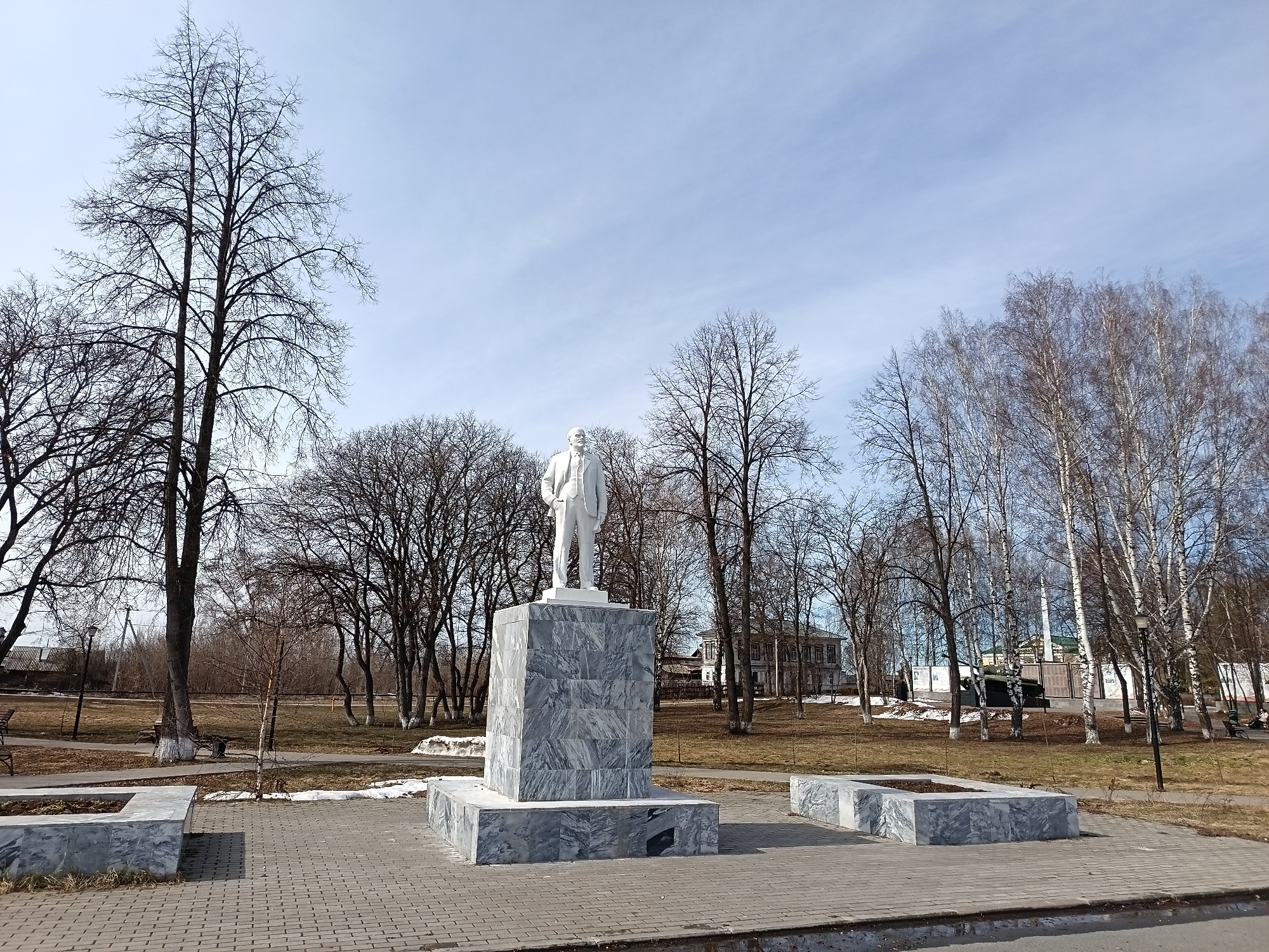 The image depicts a statue of Vladimir Lenin in the city of Osa, Russia. The statue is made of white marble and stands on a grey marble pedestal. The statue stands in a park setting with bare trees, benches, and paving stones. A few patches of snow are present on the ground. In the background, there is a fence, a war memorial with a tank, and a building. The sky is partly cloudy.
