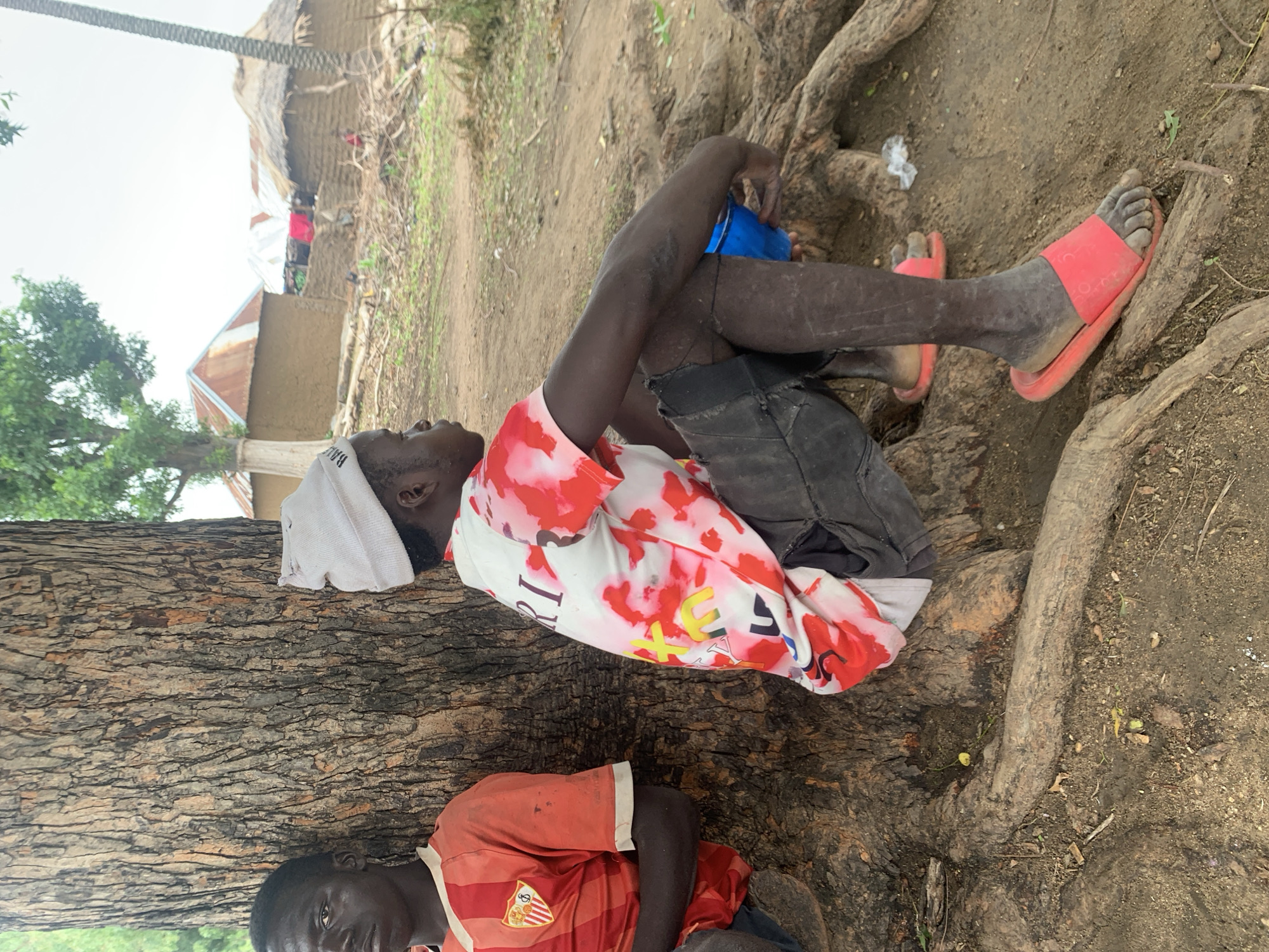 In Dull A, Nigeria, two young men are pictured sitting under a large tree. The man on the left, wearing an orange and red striped t-shirt, leans against the tree trunk. To his right, another young man, wearing a white beanie and a t-shirt with a red and white tie-dye pattern, is seated with his knees bent. He holds a blue bowl in his hand and is wearing dark shorts and red sandals. In the background, several mud-brick houses with corrugated iron or thatched roofs are visible, suggesting a rural or semi-urban environment. The ground is dirt and scattered with dry leaves and debris.