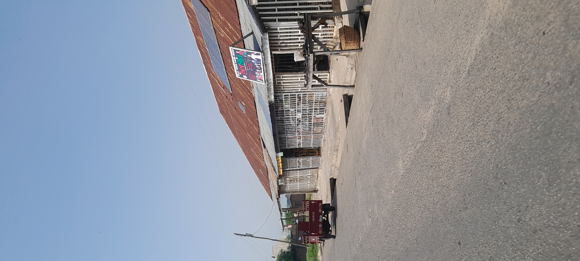 The image shows a street scene in Jalingo, Nigeria, with a focus on a row of shops lining the right side of the road. The shops have corrugated metal roofs, some of which are equipped with solar panels. The storefronts are covered with metal bars, suggesting security measures. A sign advertising "Bet9ja" is prominently displayed on the roof of one shop. In front of the shops, there is a wooden structure with a basket.
On the left side of the road, a red motorized tricycle with a cargo bed is parked. The road is paved with asphalt. A utility pole with wires stands near the tricycle. The sky is clear and blue. The environment appears to be dry and sunny, consistent with a warm climate. No people are clearly visible in the image, though there may be figures obscured within the shops or the tricycle. The overall scene depicts a typical commercial area in a Nigerian city.