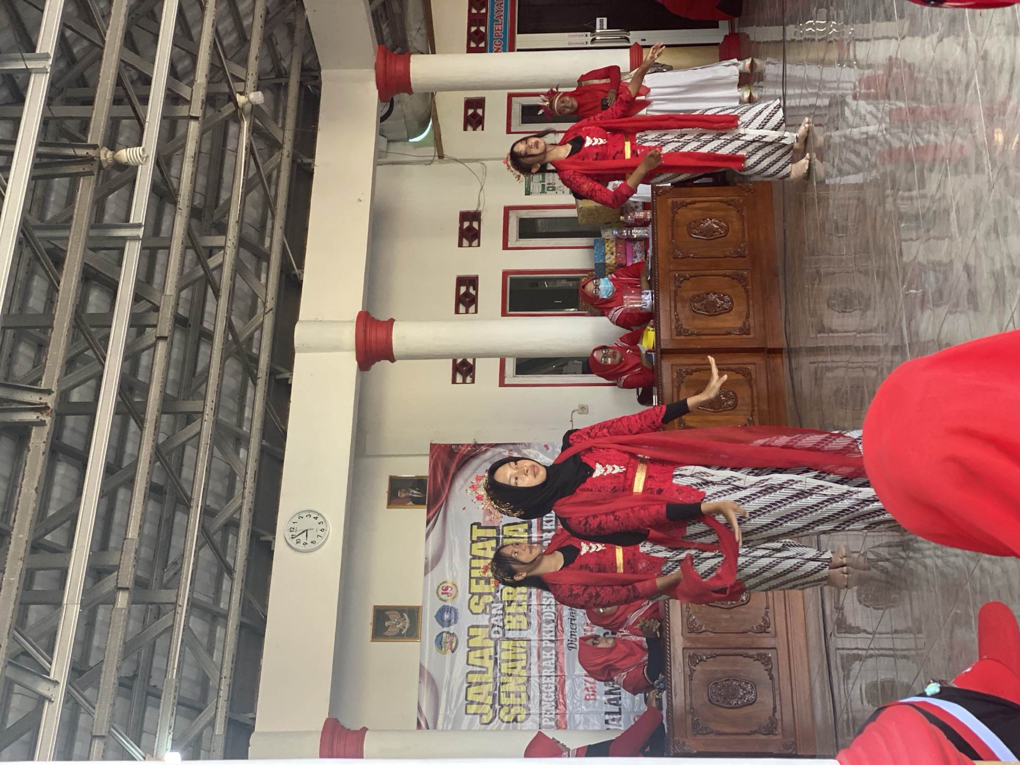 In the city of Gumelar Kidul, Indonesia, an indoor event is taking place, likely a community gathering or performance. The scene is dominated by four young women dressed in traditional red and white Indonesian attire, engaged in a dance or performance. Their outfits consist of patterned trousers, red tops with gold accents, and red shawls draped over their shoulders. Two of the dancers wear intricate headpieces, suggesting they are the main performers.
Behind the dancers, a long wooden counter or stage area is visible, behind which several other individuals are seated. Some are also wearing red headscarves and traditional clothing, suggesting they are organizers or participants. One woman behind the counter wears a face mask. A large banner behind them reads "JALAN SEHAT DAN SENAM BERSIHA" and "PENGGERAK PKK DESA" which translates to "HEALTH WALK AND CLEAN EXERCISE" and "VILLAGE PKK MOVER", indicating the event's purpose.
The environment appears to be a simple indoor hall with a high, exposed metal truss ceiling. White walls with decorative red elements and framed pictures are visible. A prominent analog clock on the wall shows the time as approximately 8:50. The floor is tiled and reflective, catching the light. The overall atmosphere suggests a vibrant cultural event within a community setting.