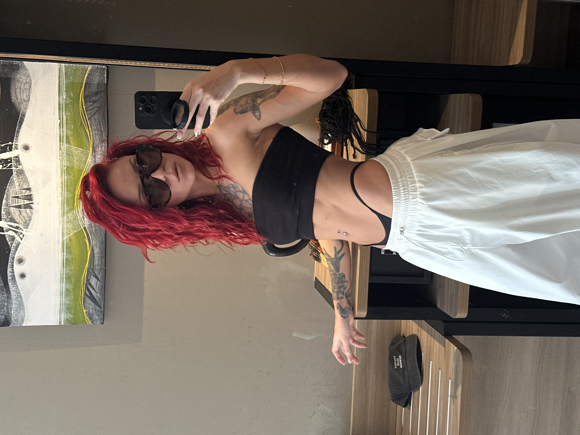 A woman with red hair and sunglasses takes a selfie in front of a mirror, wearing a black top and white pants. The background includes a painting on the wall and a wooden structure. The location is Unknown, Indonesia.