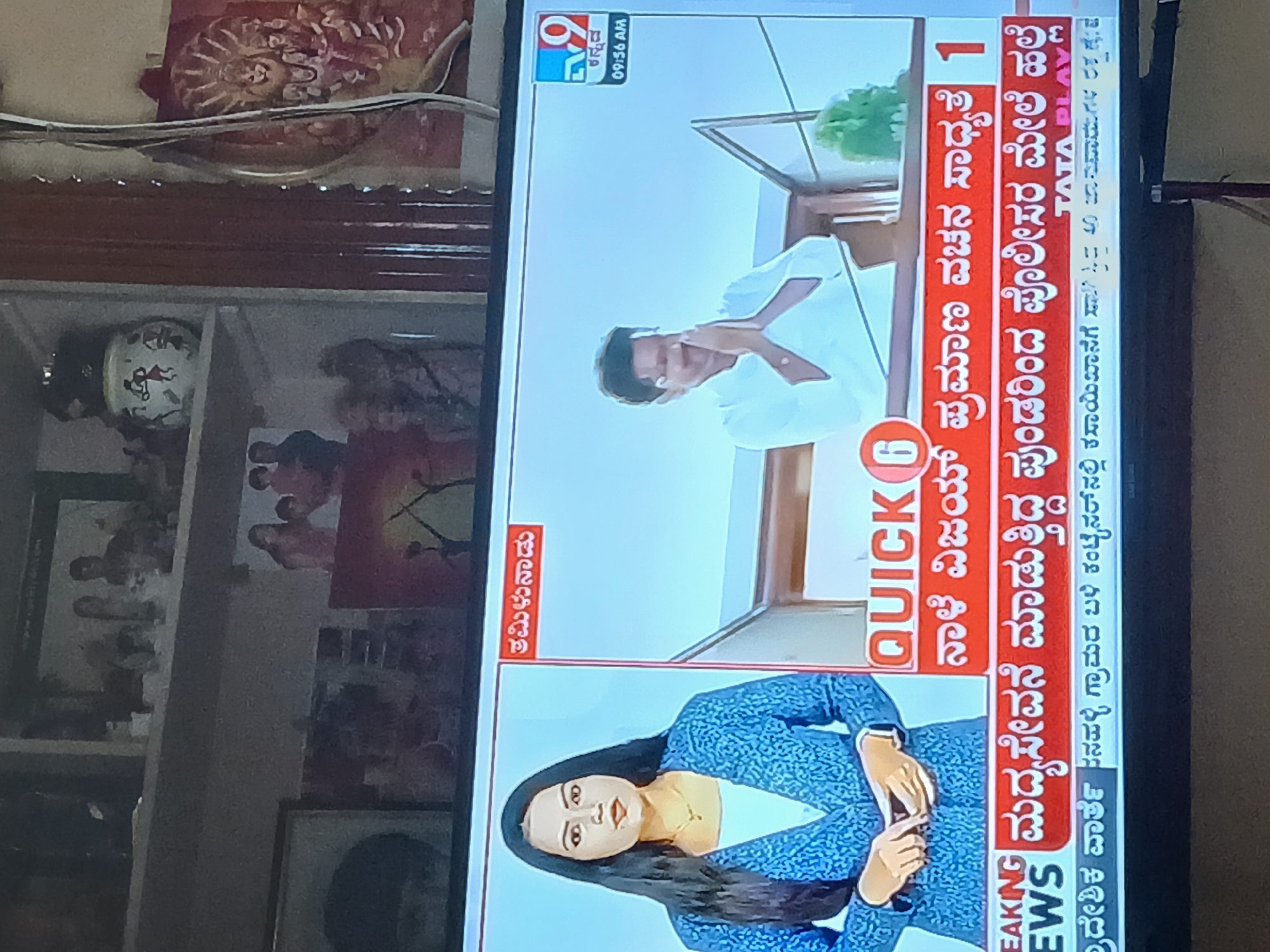 The image shows a television screen displaying Kannada news. The television is set in a room with shelves that hold items such as vases, paintings, and figurines. The news screen features an anchorwoman in a blue dress and a man in a white shirt. The screen displays text in Kannada, including the time "09:56 AM". Other details visible on the screen include the news channel logo, "TV 9," and a mention of "Quick 6." The news segments discussed involve events related to political proceedings and law enforcement. The location is in a residence in Bengaluru, India.