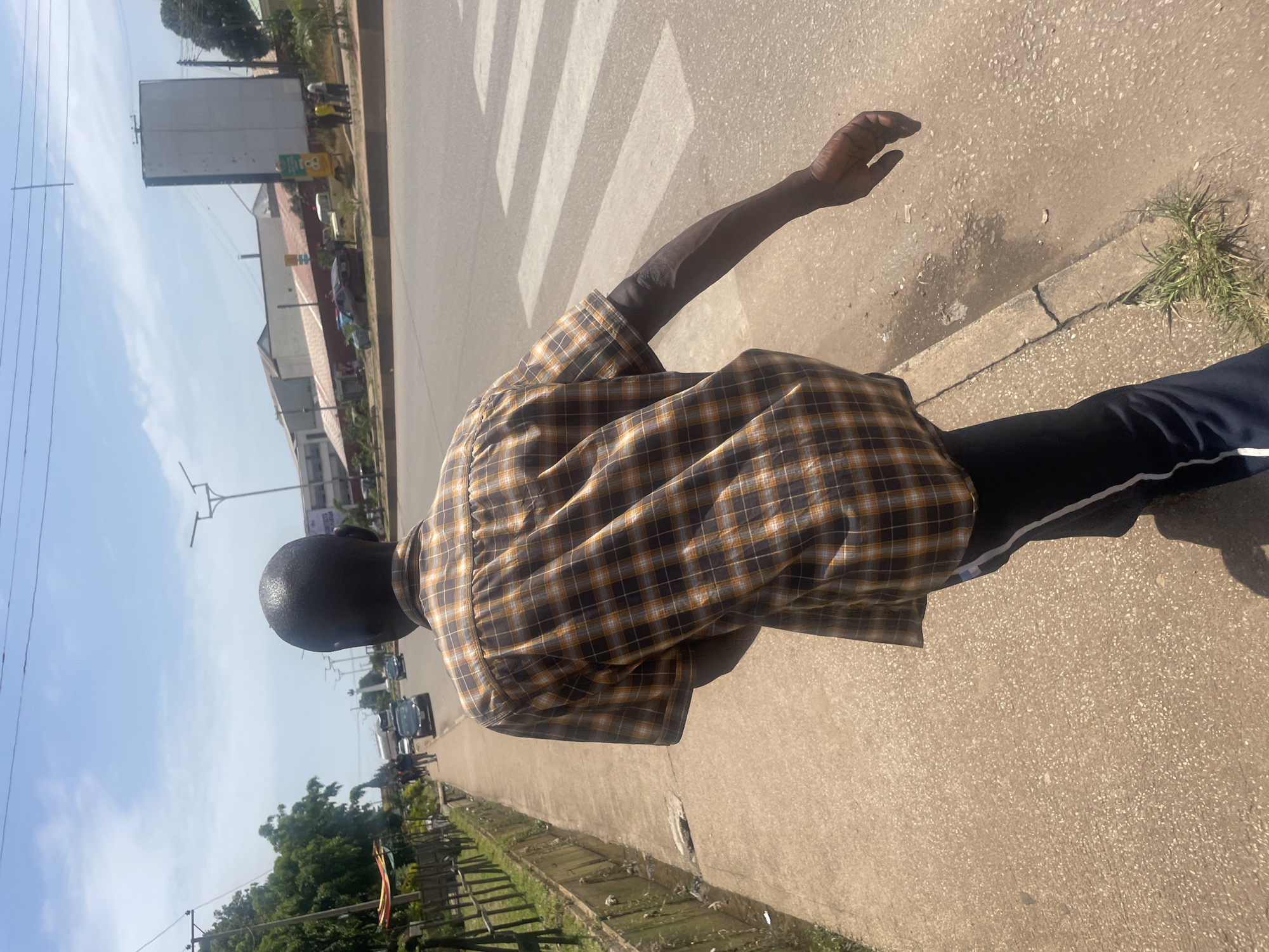 A Black man walks along a sidewalk next to a street. He is wearing a brown and orange checkered short-sleeved shirt and dark pants with a white stripe. His head is bald, and he is looking forward. The sidewalk is made of concrete, and there is a drainage ditch to the left. Across the street, there are buildings, trees, and vehicles. A pedestrian crossing is visible on the street. The sky is blue with some clouds. The scene appears to be in a city or town.