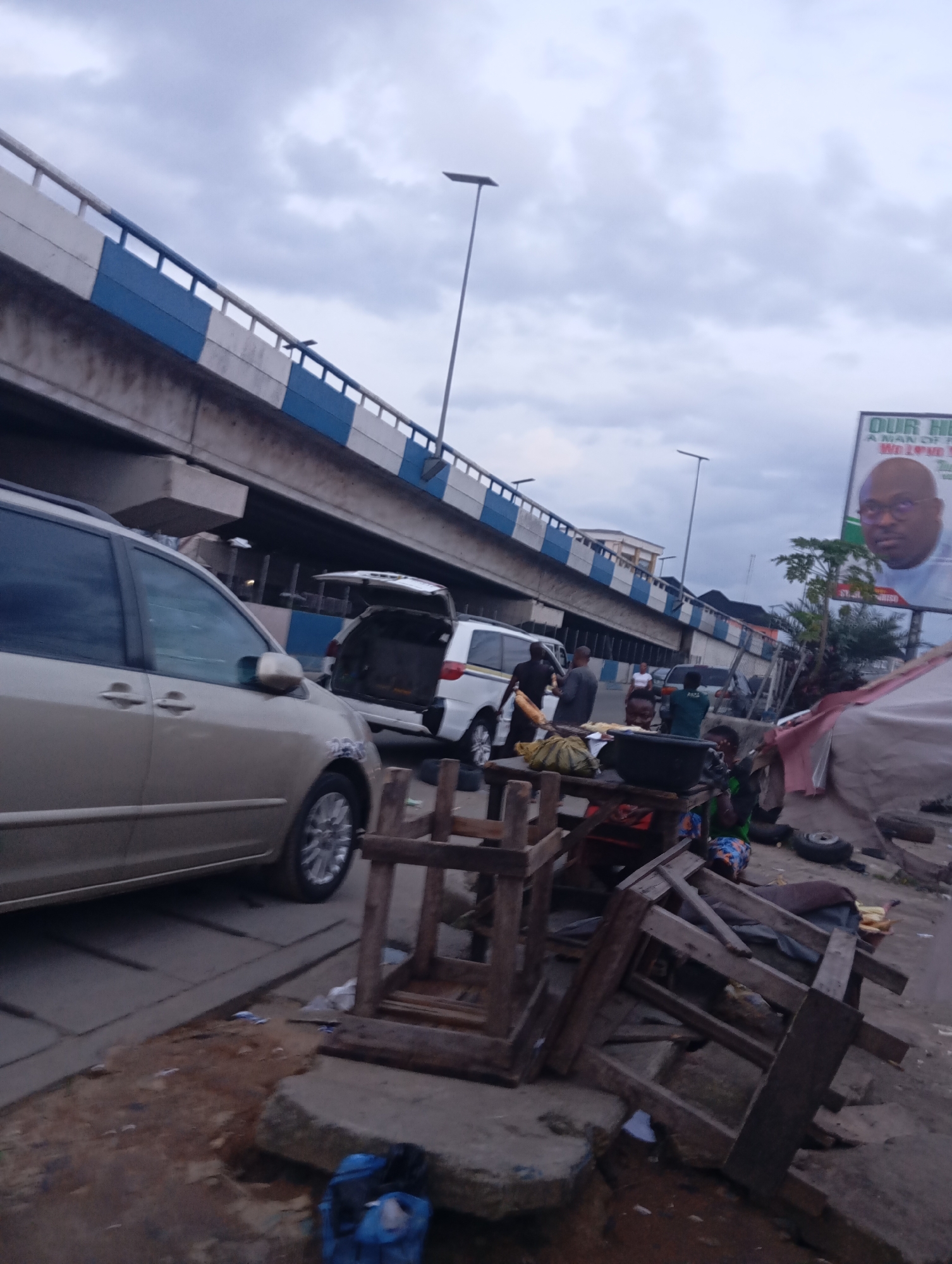 The image depicts an outdoor scene in Port Harcourt, Nigeria, likely a busy roadside environment under an elevated highway. In the foreground, several dilapidated wooden chairs are stacked haphazardly on a patch of uneven, muddy ground. A blue backpack rests on a concrete slab to the left. A light-colored minivan is parked in the middle ground, with its rear hatch open. Behind it, another white minivan is visible. To the right, a large billboard featuring a man's portrait with the text "OUR HE..." and "A MAN OF F..." is partially obscured by a makeshift stall covered with a reddish-brown tarp. Several people are present, some appearing to be vendors operating from small tables, with produce and containers visible. The sky is overcast with heavy clouds. The elevated highway structure dominates the upper portion of the image, with white and blue markings along its side. The overall scene suggests a dynamic, possibly informal, commercial area integrated with urban infrastructure.