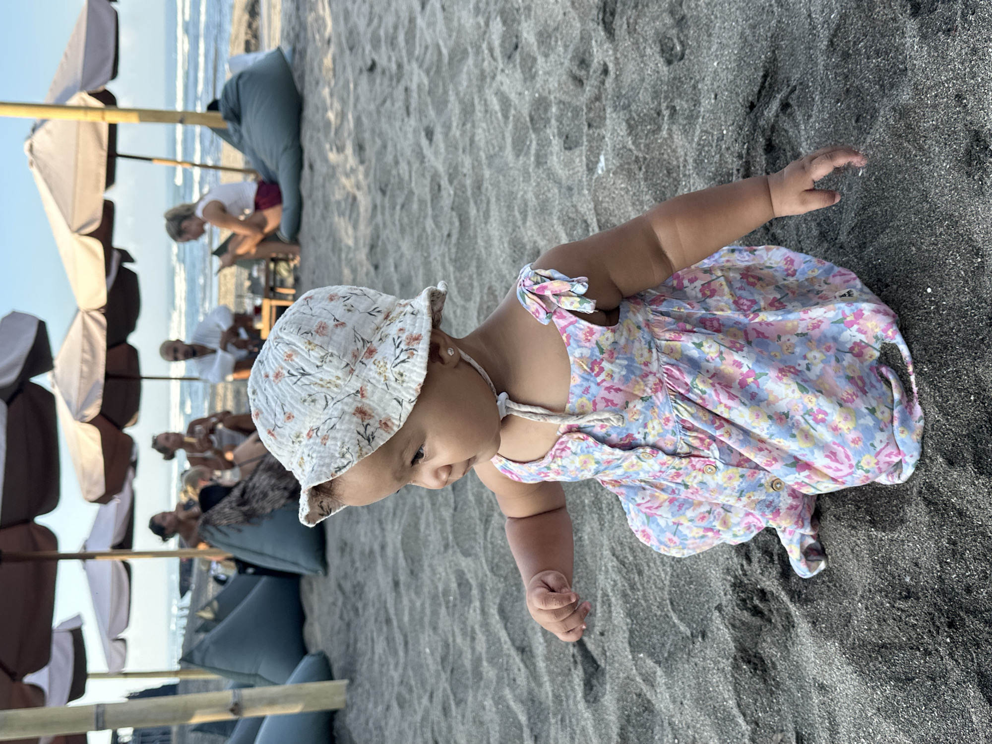A young child wearing a floral dress and a bucket hat is crouched on a black sand beach in North Kuta, Indonesia. The child is facing to the left, with their right hand outstretched. In the background, several people are seated under umbrellas, and the ocean is visible. Beanbag chairs are scattered on the sand. The sky is clear and blue.