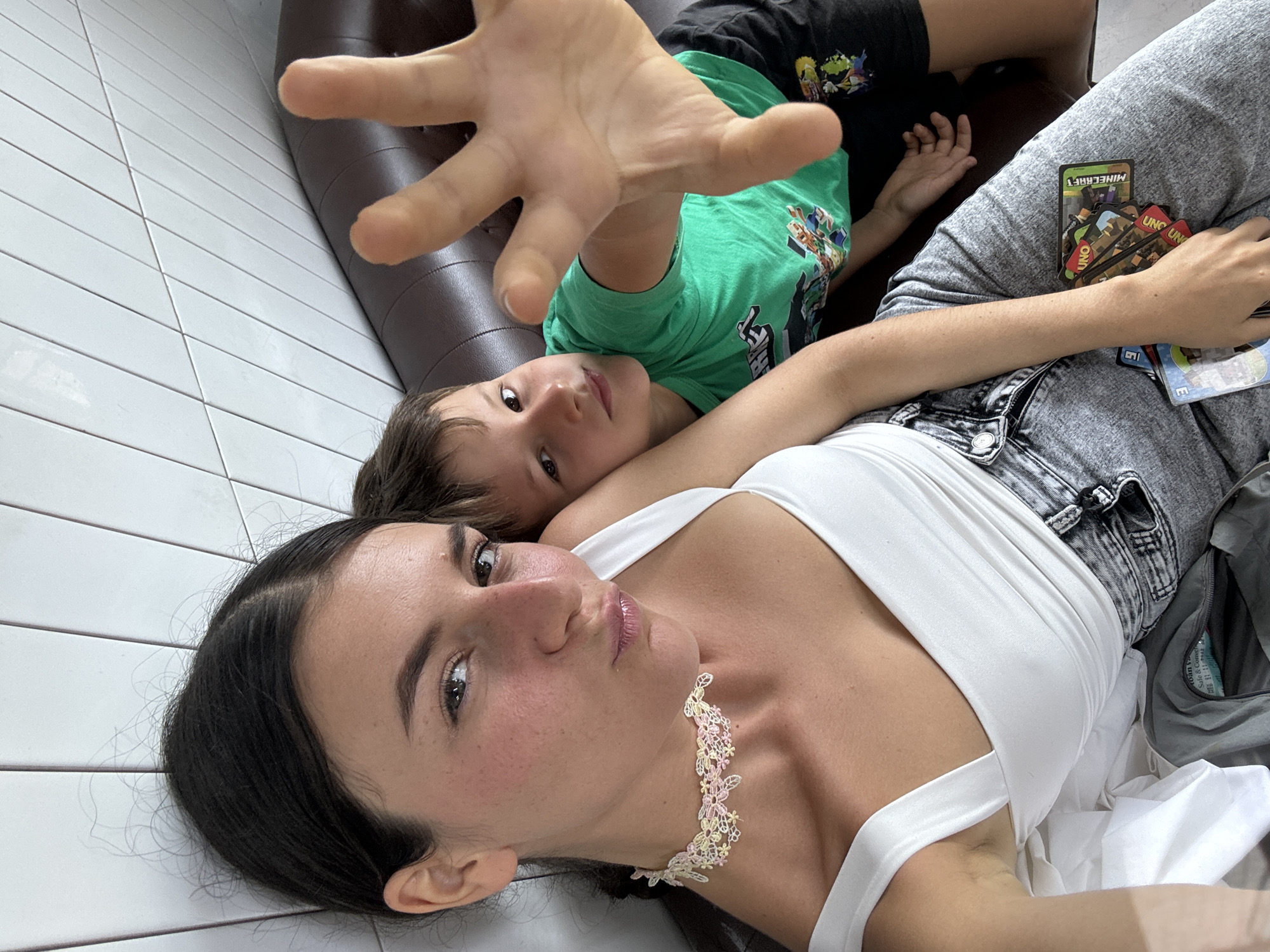 The image shows two individuals, a woman and a boy, seated on a brown leather sofa. The woman, in the foreground, is looking at the camera with a playful expression, her lips pursed. She is wearing a white top and a delicate lace choker. The boy, partially behind her, is also looking towards the camera, with his right hand extended forward, obscuring part of the frame. He is wearing a green t-shirt and dark shorts. On the woman's lap, several UNO cards are visible. The background consists of a wall tiled with rectangular white tiles. The lighting suggests an indoor setting. There is no indication of the specific location within Phan Thiet, Vietnam.