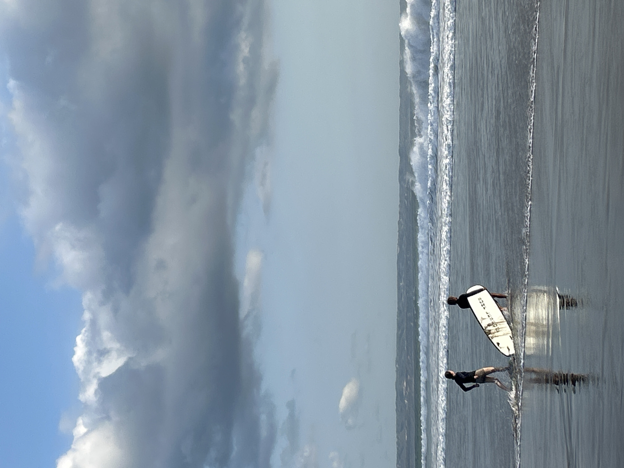 The image depicts two individuals walking into the ocean at North Kuta, Indonesia. The person in front, to the left, is wearing a dark long-sleeved top and light-colored shorts, with their body angled towards the sea. To their right, slightly behind and further into the water, a second person is carrying a white surfboard, also facing the ocean. The surfboard has dark markings on its surface. They are walking through shallow water where small waves are breaking around their legs. The ocean exhibits white-capped waves in the mid-ground. The sky above is a mix of clear blue and large, dark, dramatic clouds, suggesting a potentially overcast day or a change in weather. The overall scene is a beach environment with the ocean and sky as prominent features.