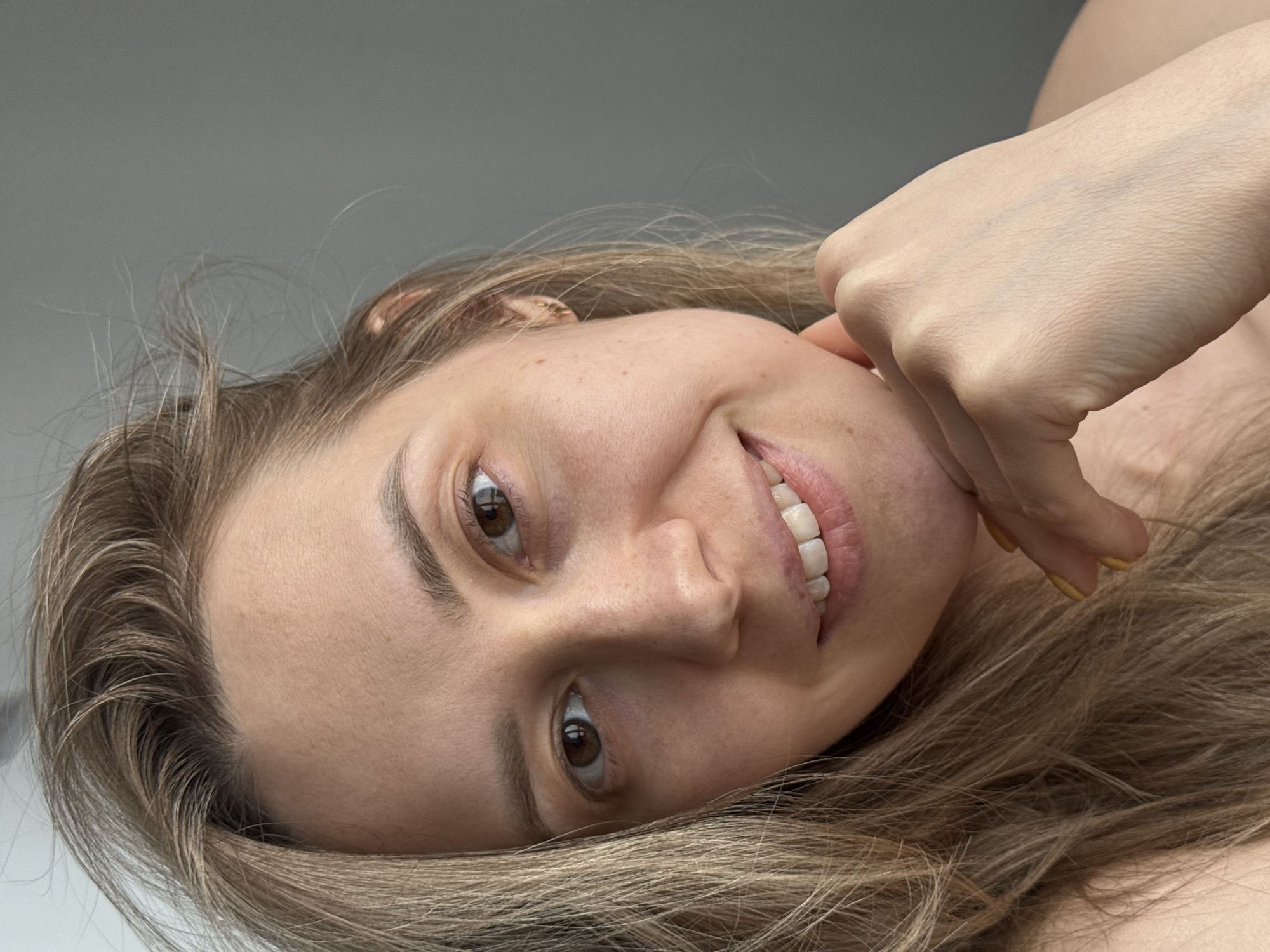 A close-up, slightly angled selfie depicts a young woman with fair skin, light brown hair, and brown eyes. She is smiling slightly, with her teeth partially visible. Her right hand is positioned near her cheek, with fingers gently touching her face. The background appears to be a plain, light gray surface. There is no information to suggest that the image was taken in Chaikovsky, Russia.