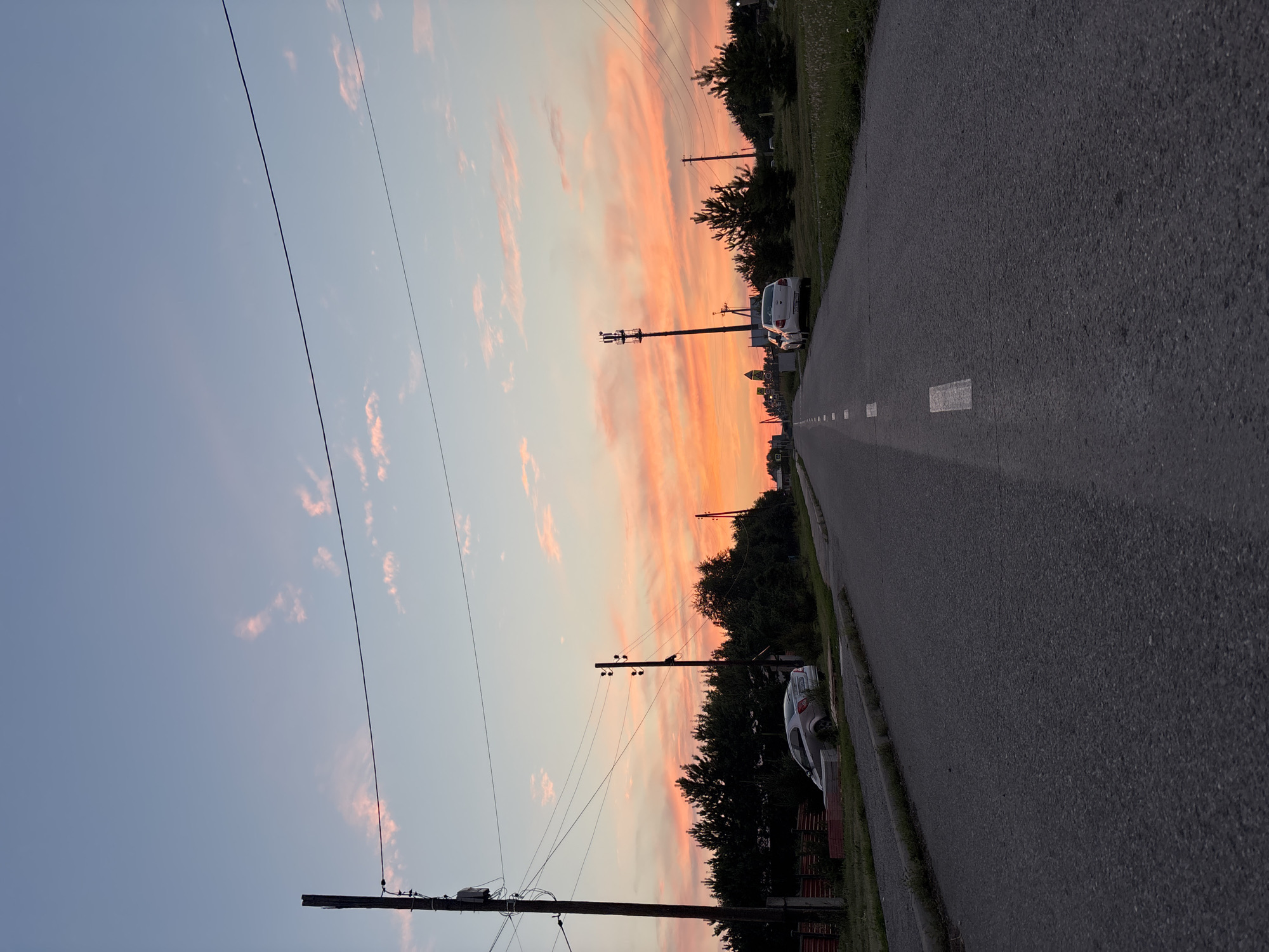 The image depicts a rural road in Vishnevka, Russia, at dusk. The sky is a gradient of pale blue at the top, transitioning to vibrant hues of orange and pink clouds closer to the horizon, suggesting a sunset. Overhead power lines stretch across the sky, supported by wooden poles. The road is paved and has a dashed white line marking the center.
On the left side of the road, a silver car is parked partially on the grass, near a wooden fence. Behind the car, a dense line of dark green coniferous trees forms a backdrop. Another wooden pole with electrical insulators stands near the car.
On the right side of the road, several young pine trees are visible, interspersed with grass. Further down the road, another white car is parked. In the distance, a cell tower and more power poles are apparent. The overall environment suggests a quiet, semi-rural setting experiencing the end of daylight. There are no people visible in the image.