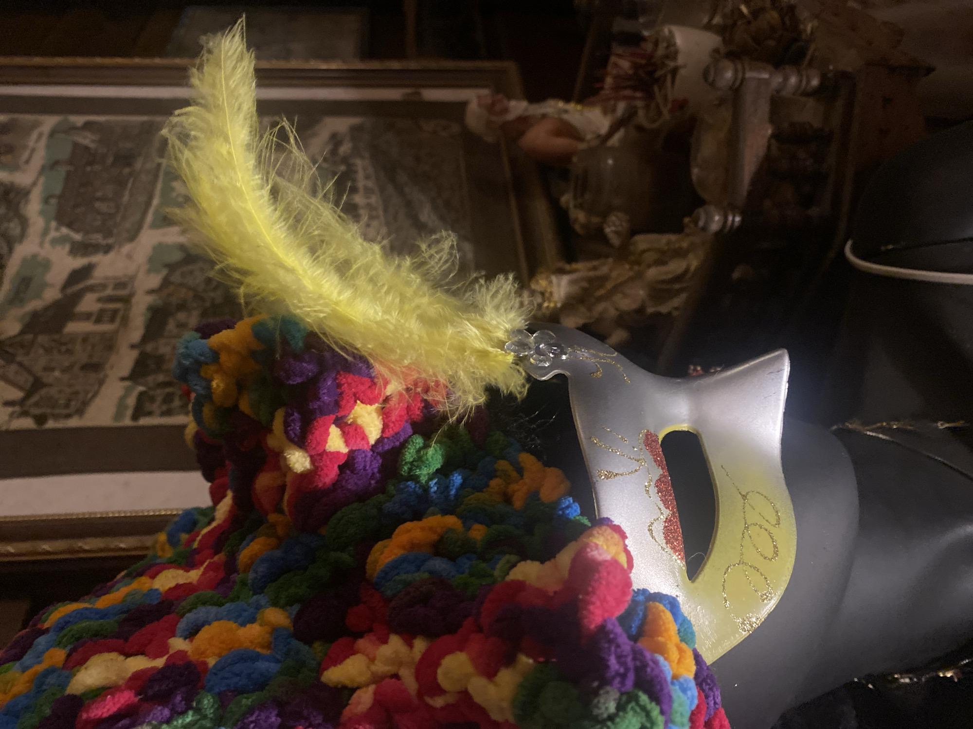 A photograph shows a silver mask with a yellow feather lying on top of a colorful crocheted item. The mask has glittery gold and red designs. In the background, there is an unclear picture in a golden frame and other decorative items. The location is difficult to determine from this image, and there is no evidence to suggest it is in Baan Don Sai, Thailand.