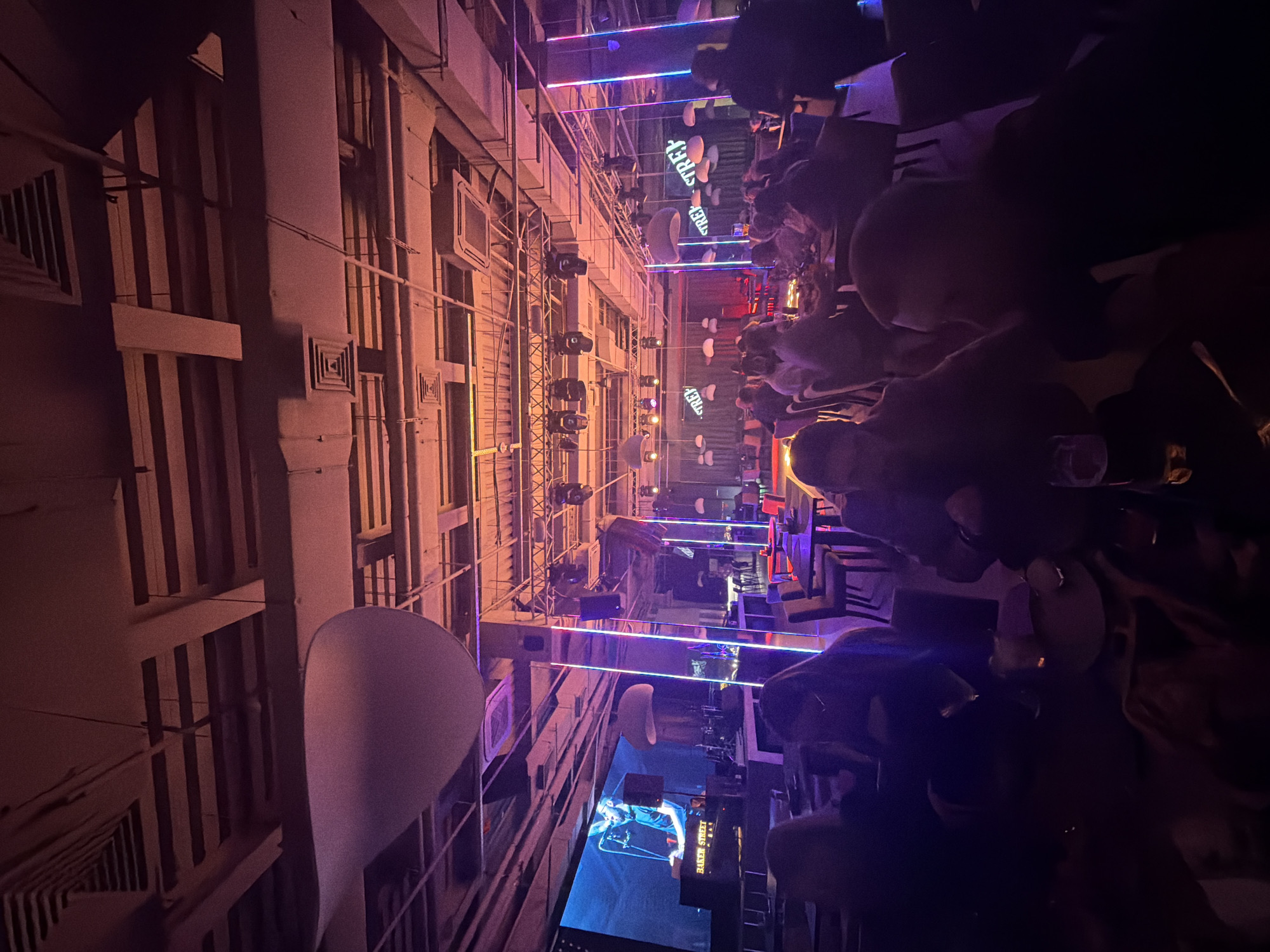 This image depicts an indoor entertainment venue, likely a bar or club in Krasnoyarsk, Russia, designed with an urban, street-themed aesthetic. The scene is dimly lit, suggesting evening or night, and features a long, narrow space resembling an alleyway or indoor street.
Numerous people are seated at tables and booths along the left side of the "street," mostly appearing as dark silhouettes enjoying the ambiance. The right side features a detailed facade mimicking a building wall, complete with window-like structures, square vents, and pipes, all bathed in a warm, reddish-orange glow from spotlights.
The central space is illuminated by horizontal strips of neon lighting in vibrant blue, pink, and purple, contributing to a modern, lively atmosphere. Several spotlights and other light fixtures are mounted on exposed scaffolding or trusses. Large, abstract, rounded white/grey shapes, possibly decorative elements like stylized clouds or oversized objects, hang throughout the space, including a prominent, spoon-shaped object in the foreground.
In the background, at the far end of the "street," a large screen displays an image of a person, with the text "Elena Shebert" visible below them, suggesting live entertainment or a video presentation. Multiple glowing signs with the word "STREEF" are also visible along the venue's length. The overall impression is a bustling, social environment designed for leisure and entertainment.