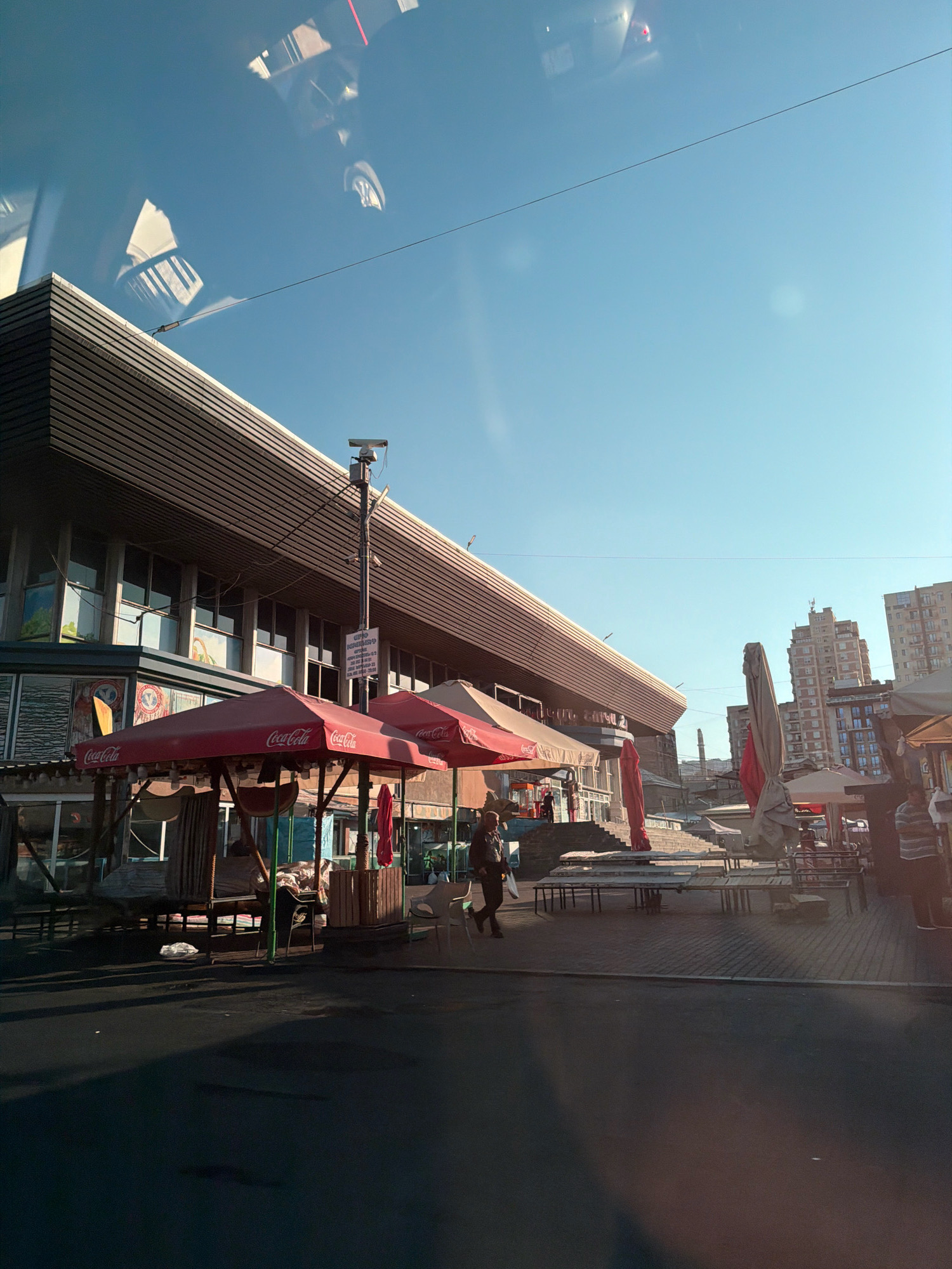 The image shows a street scene with market stalls under red Coca-Cola branded umbrellas. A large, modern building with a slatted facade dominates the left side of the frame. The building appears to be a market or commercial complex. Several people are visible in the scene, including one person walking past the stalls, another sitting at one of the stalls, and a few others in the background near the complex. The ground is paved with cobblestones. In the background, tall residential buildings are visible, suggesting an urban environment. The sky is clear and blue, with some reflections visible on what appears to be a glass surface in the foreground, possibly a window or a car windshield. The overall impression is that of a bustling open-air market on a sunny day in Yerevan, Armenia, likely near a large market complex.