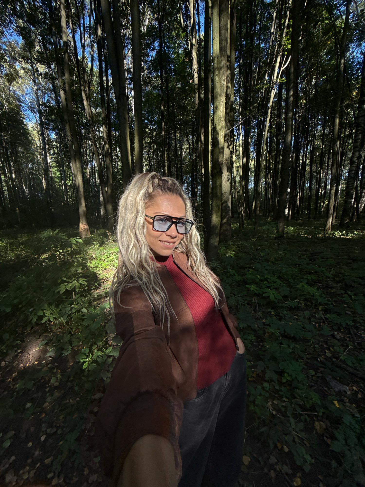 A blonde woman with a high ponytail is taking a selfie in a sun-dappled forest. She is smiling at the camera, wearing a red top, a brown jacket, and dark jeans. She is also wearing large, dark-rimmed glasses. The forest floor is covered in green foliage and fallen leaves, and tall trees with dark bark surround her. The lighting suggests it is daytime. The scene is located in the city of Rusanovo, Russia.