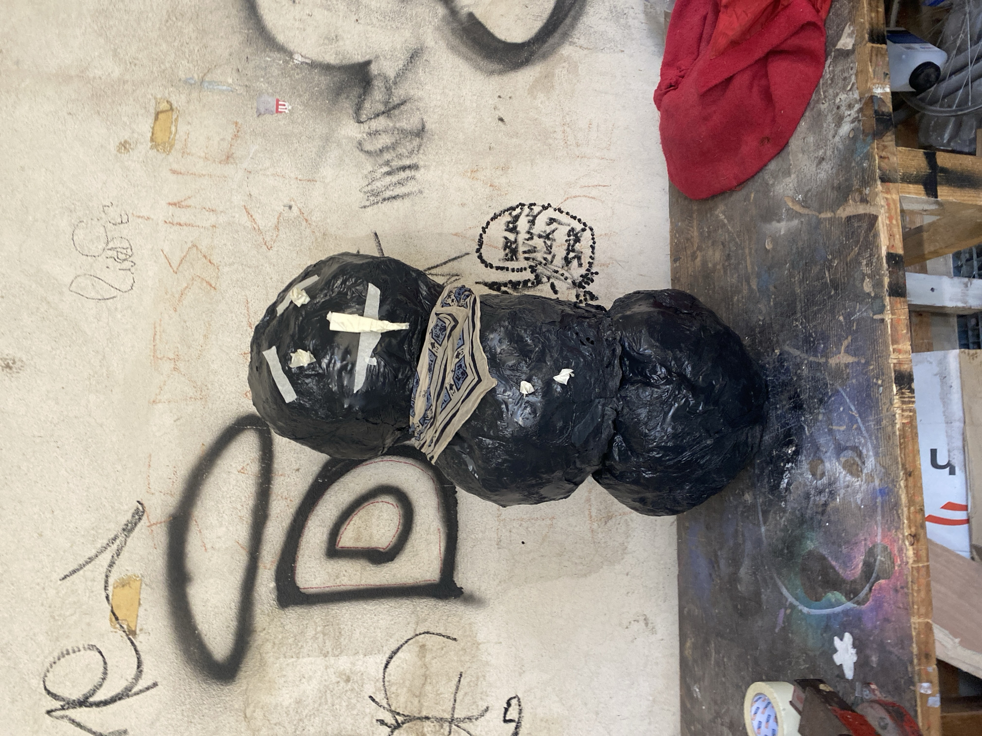 The image shows a close-up view of three black spherical objects taped together on a wall with graffiti and a wooden table nearby. The wall has various black graffiti markings, including circles, lines, and some illegible writing. There is also some faded orange or brown writing. The spherical objects are wrapped in black plastic and held together with tape. One object has a patterned cloth draped over it. A red cloth sits on the wooden table. The table is scratched and marked, with hints of colorful paint or residue. Other items are visible on the table and behind the table, including a tape roll and a white box. It is not possible to determine the location in the city of Paris from the image. There are no people visible in the image.