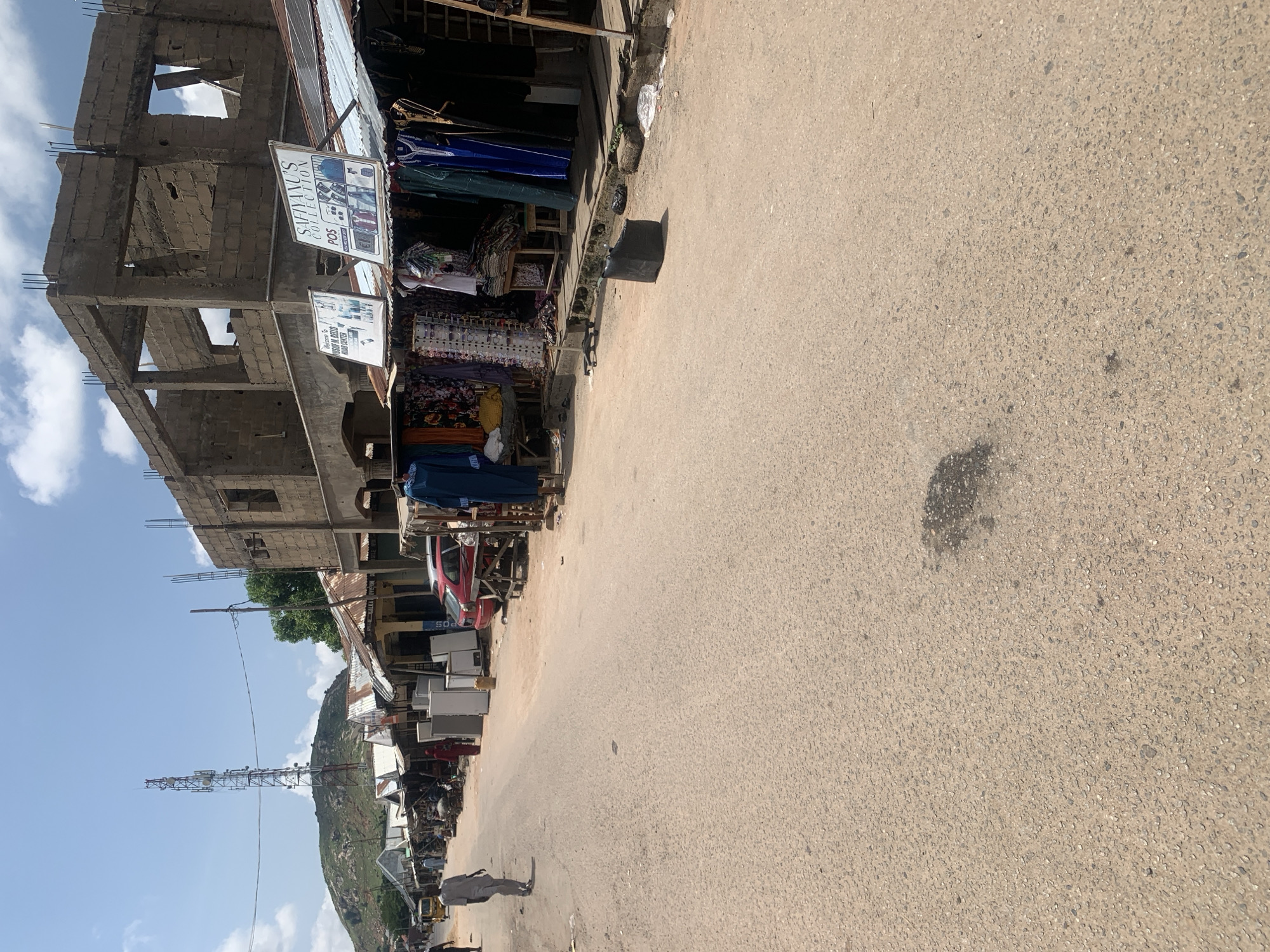 The image depicts a street scene in Jalingo, Nigeria, likely a market area. A paved road runs through the center, with stalls and shops lining the sides. Some of these structures appear to be under construction, with exposed brickwork and reinforcement bars visible. One building features a prominent cell tower behind it, situated on a grassy hill. Various goods are displayed for sale in the stalls, including clothing, refrigerators, and other items. Several people are present, walking along the road or attending to the shops. A red car is parked near one of the stalls. The overall environment suggests a busy commercial district with elements of ongoing development.