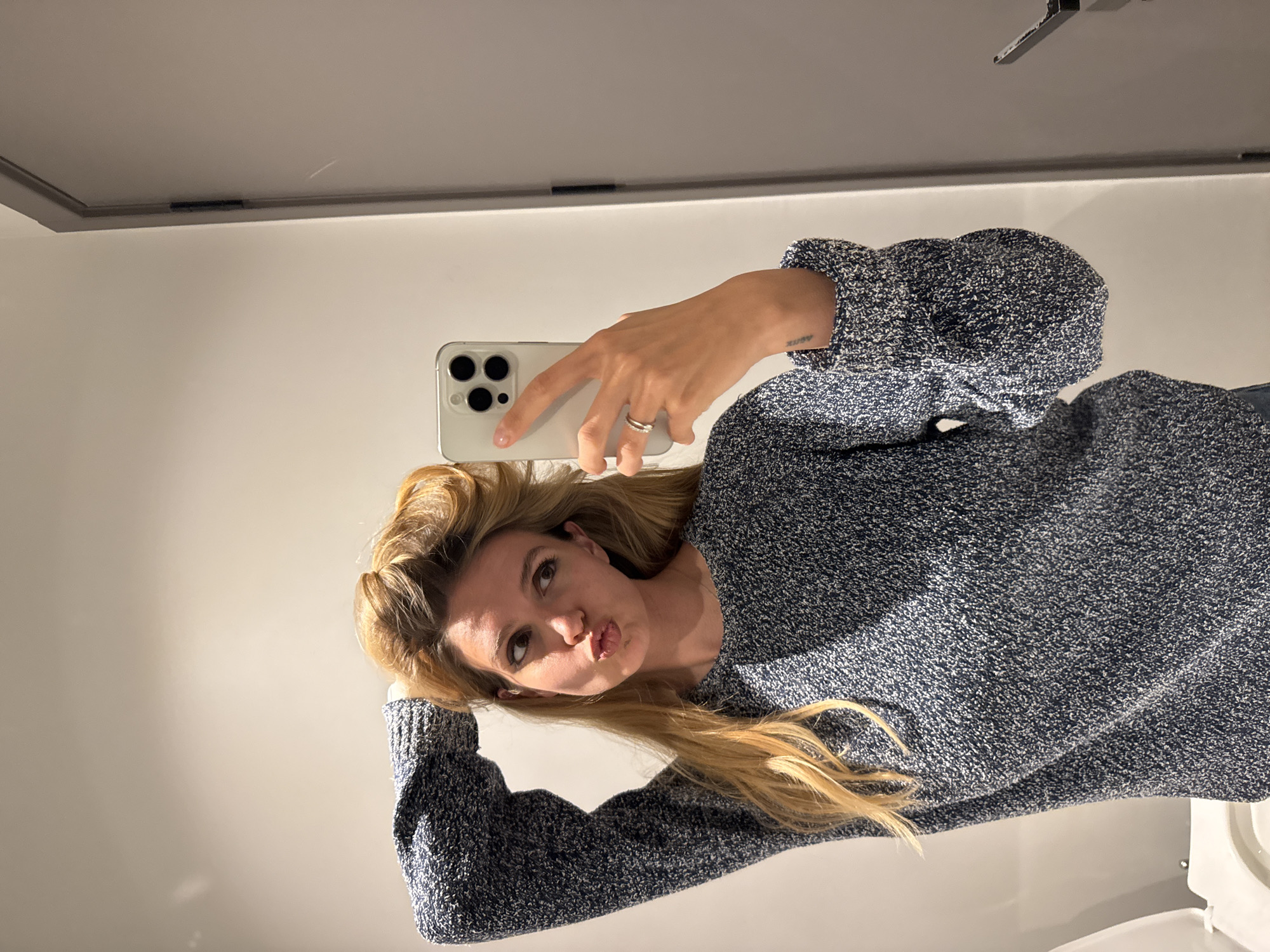 A woman with blonde hair is taking a selfie in a bathroom. She is wearing a blue and white speckled sweater. She is holding a white smartphone in her right hand. Her left hand is resting on her head. She is puckering her lips. The bathroom has white walls and a white ceiling. A small portion of the toilet is visible in the lower right corner. Location: Unknown, Unknown.