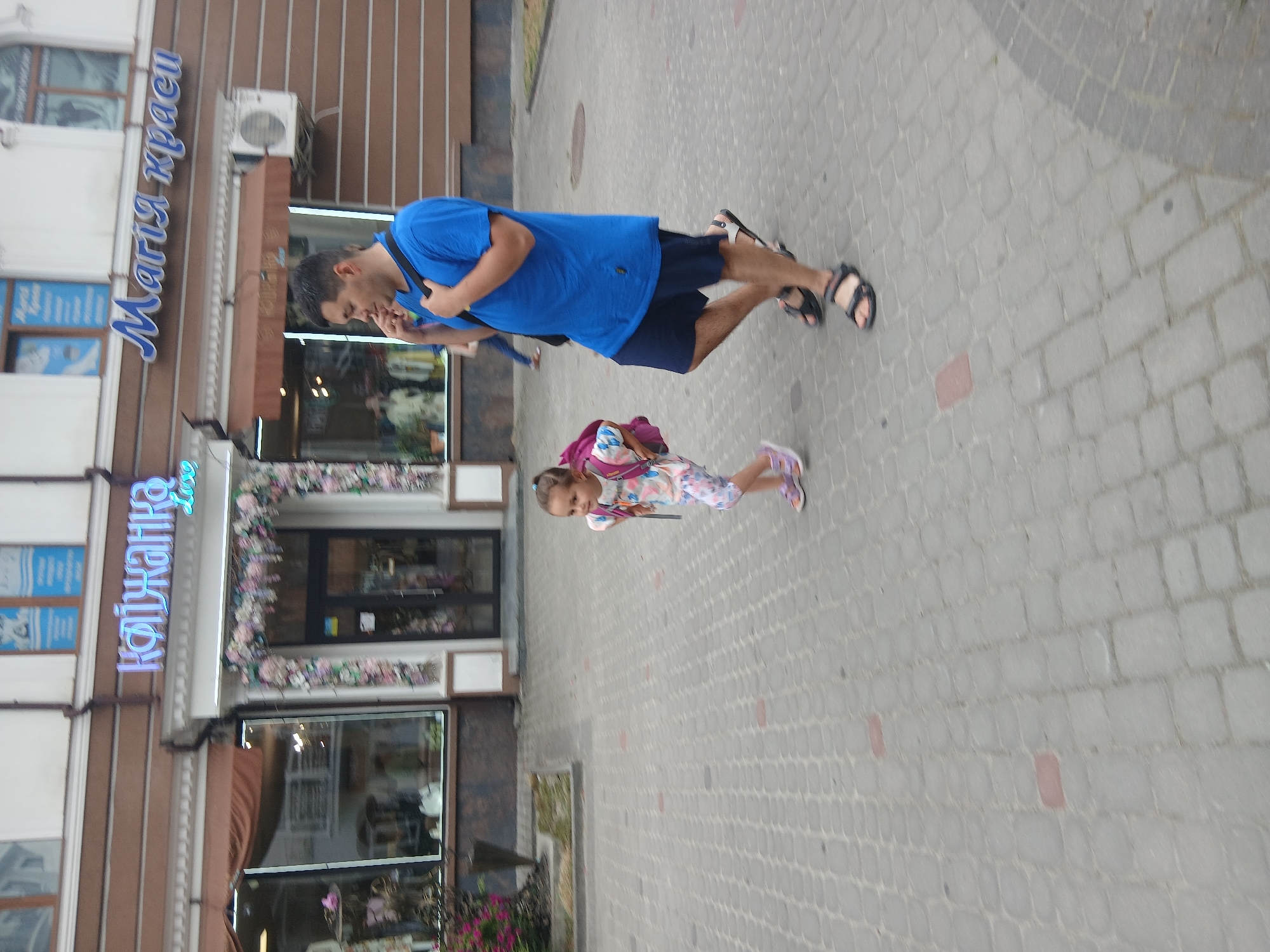 The image captures a street scene in Truskavets, Ukraine. Two people are visible: a man wearing a blue t-shirt and dark shorts, and a young girl in a colorful dress and pink sandals. The man is walking, his head turned downwards as if looking at something. The girl, with a backpack, is also walking. They appear to be on a paved sidewalk in front of a building with storefronts.
One storefront has a blue neon sign with Cyrillic text that reads "Марія краси" (Maria krasy), which translates to "Maria Beauty". Another sign, also in blue neon, reads "Квіжанка" (Kvizhanka), which could be a shop name. The building has a modern facade with windows and vertical brown paneling. Flowers are visible decorating the area above the storefront windows. An air conditioning unit is mounted on the building.
The scene suggests daytime, with ambient light illuminating the area. The ground is paved with cobblestones. The overall atmosphere appears to be one of casual outdoor activity on a street or promenade.