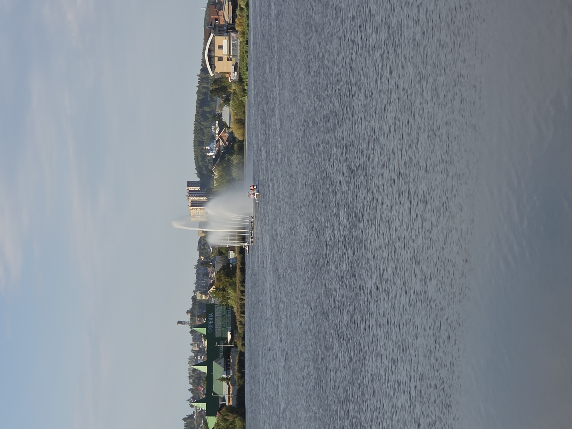 The image shows a wide, calm body of water with a city skyline in the background. In the foreground, the water is dark blue and rippled, reflecting the sky. In the middle ground, there is a large fountain spraying water high into the air. To the left of the fountain, a small pedal boat with two people is visible. The city skyline features a variety of buildings, including a large green building with pointed roofs and the word "ОРБИТА" (Orbita) in Cyrillic letters, a tall, slender building, and several smaller residential and commercial buildings. A church with a blue dome is also visible. The background is a tree-covered hill. The sky is a pale blue with some wispy white clouds. The scene appears to be a waterfront park or recreational area in the city of Miass, Russia. The lighting suggests it is late afternoon or early evening.