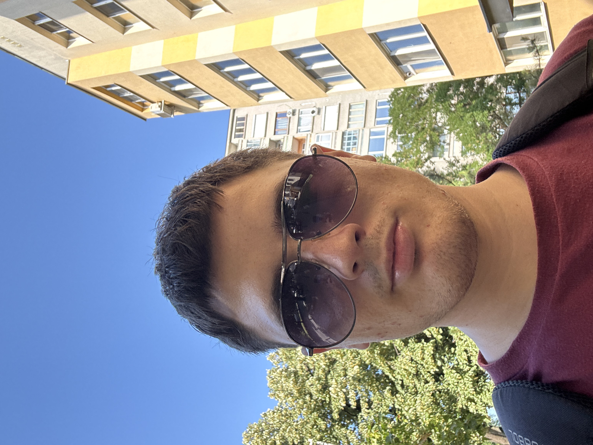 A selfie taken outdoors shows a young Caucasian man wearing a maroon t-shirt and dark aviator sunglasses. He has short brown hair and is looking directly at the camera. Behind him, a bright yellow apartment building stands tall against a clear blue sky. To the left of the building, lush green trees are visible, suggesting a residential area. The scene appears to be captured on a sunny day. The building style is characteristic of residential blocks found in Stara Zagora, Bulgaria.