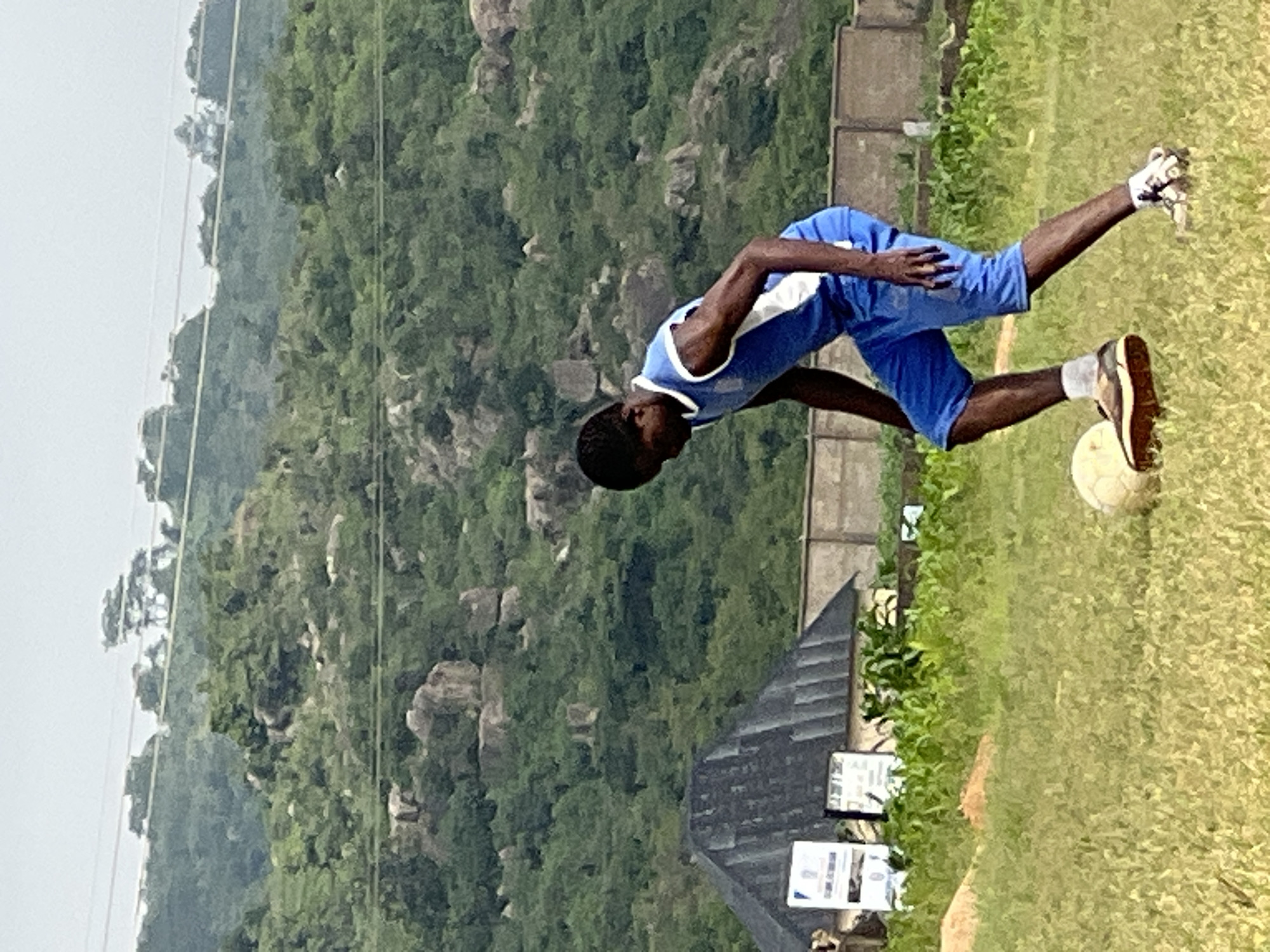 A young Black male, appearing to be a teenager, is playing soccer on a grassy field in a rural or semi-rural area. He is wearing a blue athletic uniform with white trim, consisting of a sleeveless jersey and shorts, and white socks with athletic shoes. The boy is in mid-action, dribbling a worn, off-white soccer ball with his right foot. His body is angled downwards, focused on the ball.
The background features a gently sloping, rocky hillside covered with dense green vegetation. Several large rocks are visible on the hillside. In the mid-ground, a light-colored wall runs horizontally across the frame, with a dark, sloped roof structure visible behind it, possibly a building with a corrugated metal roof. Some green, leafy plants, possibly corn stalks, are growing in front of the wall. Signs are also visible behind the boy and to the left, mounted on poles. The sky is overcast and light gray. The overall impression is a casual sporting activity in a natural, somewhat undeveloped environment. The scene is likely in or near Jos, Nigeria, given the terrain and vegetation which are characteristic of the Jos Plateau region.