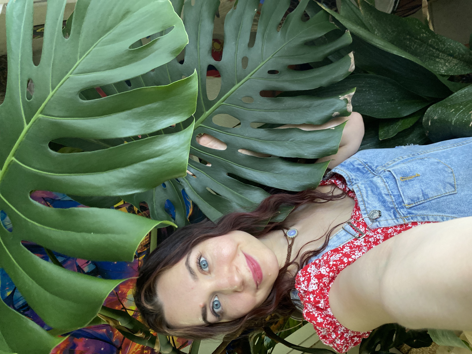 In this media file, a young woman with long, dark, wavy hair and striking blue eyes is the central subject. She is lying down, her head resting on something unseen, and looking directly at the camera with a gentle smile. She is wearing a denim jacket over a red dress with a white floral pattern. A necklace with a light-colored pendant rests on her chest.
The setting appears to be outdoors, amidst lush greenery. Large, distinctive monstera leaves frame the shot, creating a natural and vibrant backdrop. The background also shows other large, dark green leaves, suggesting a heavily planted area, possibly a garden or a room filled with many plants. The lighting indicates it could be daytime, with natural light illuminating the scene. The overall mood is relaxed and cheerful. There is no visible text in the image.