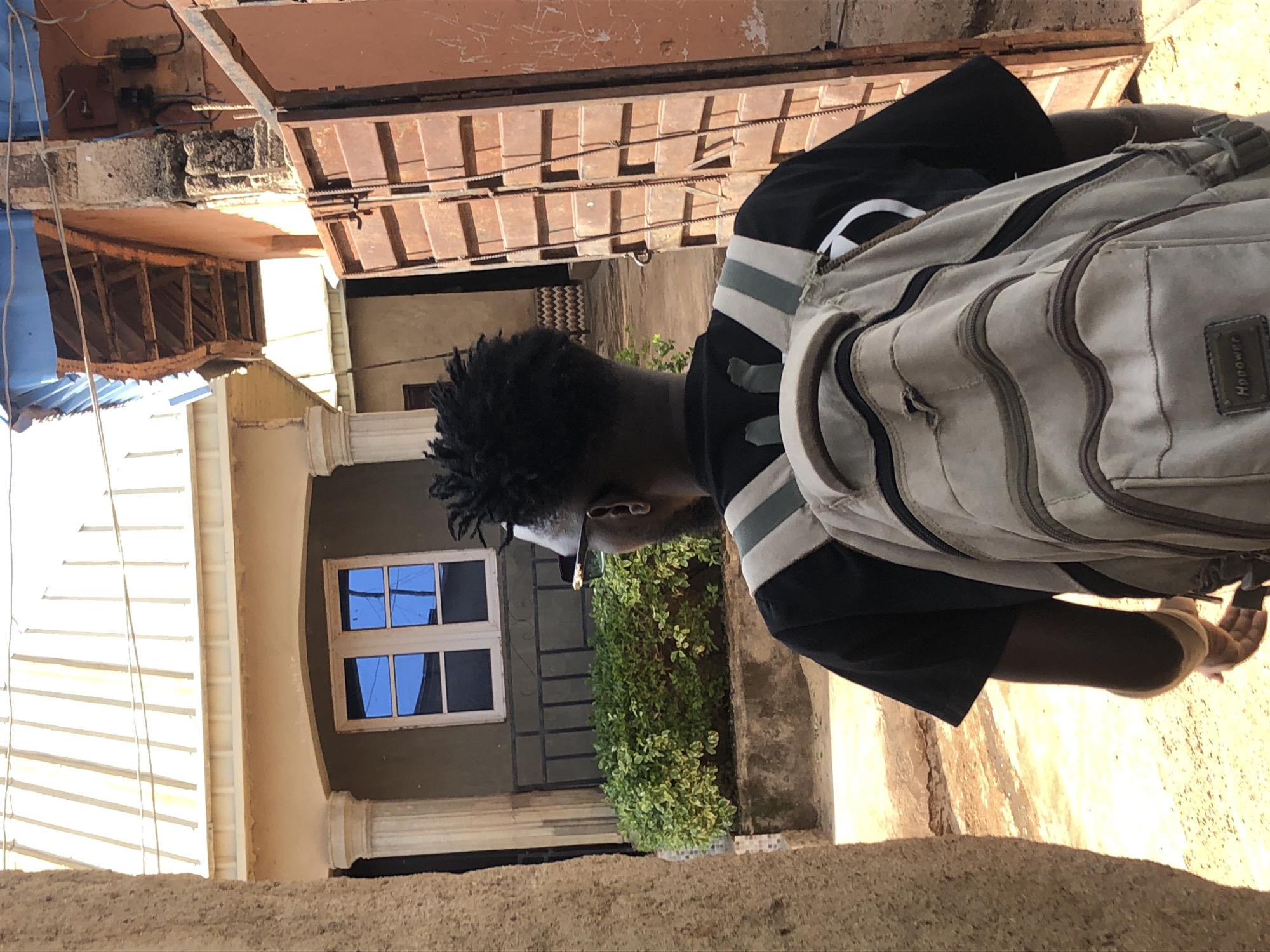 A person wearing a black t-shirt and a tan backpack walks away from the camera down a narrow, sun-drenched path. The person has dark, short, dreadlocked hair and wears sunglasses. To the left of the path, there is a light-colored building with a white tiled roof, white pillars, and a window with blue-tinted glass. A dense green hedge is planted in front of the building, partially obscuring a black metal railing. To the right of the path, there is a large, rusty, orange metal gate that is partially ajar, revealing a section of a weathered, orange-walled building behind it. Overhead, a power line stretches across the scene. The ground is covered in light brown dirt and gravel.