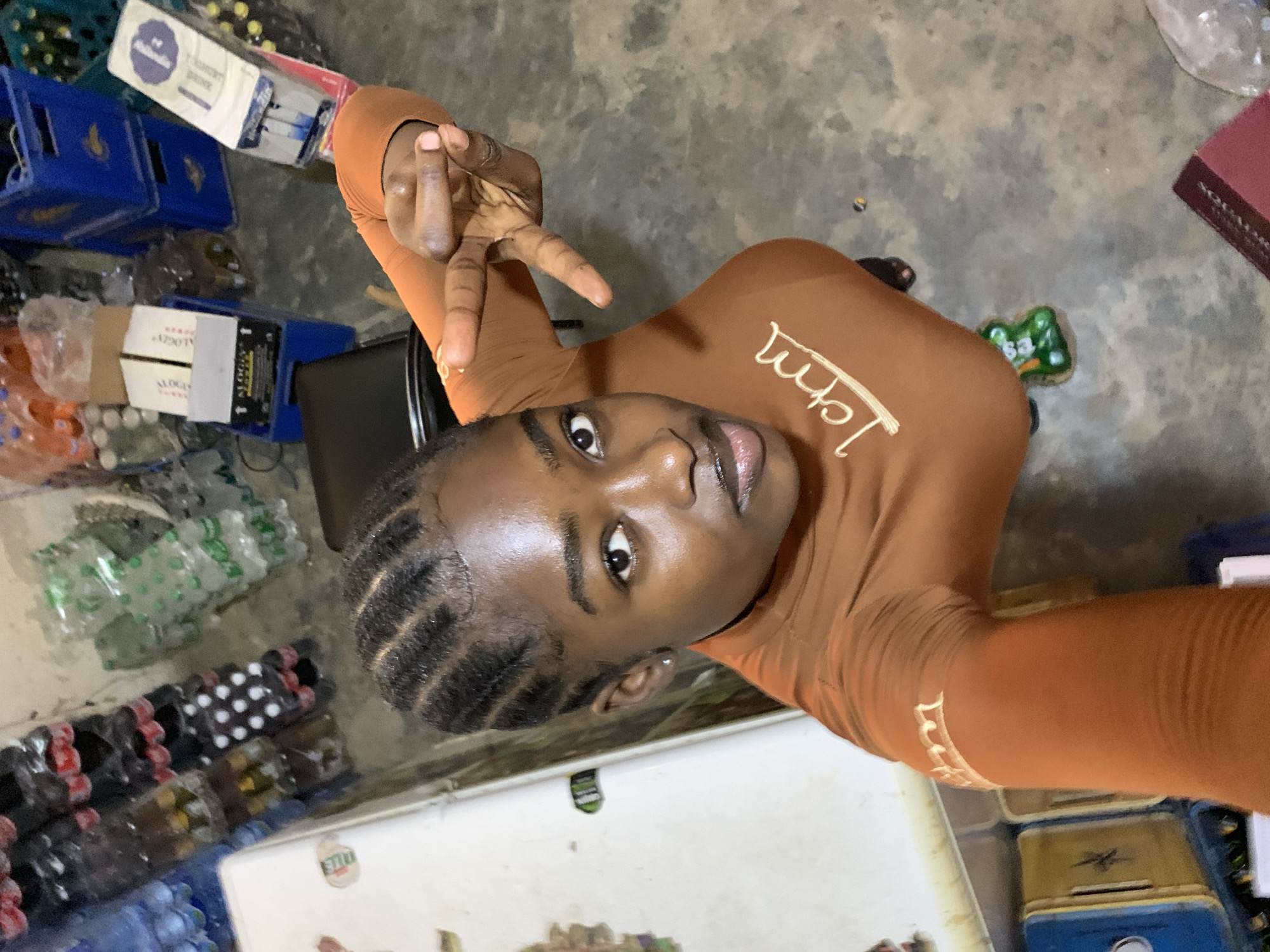 A young woman with braided hair, wearing a brown long-sleeved shirt with white lettering on the sleeve, is posing for a selfie while holding up two fingers. The setting appears to be a small store or storage room with stacks of bottled beverages, crates of drinks, and boxes lining the floor. The location is identified as New Stadium, Nigeria.