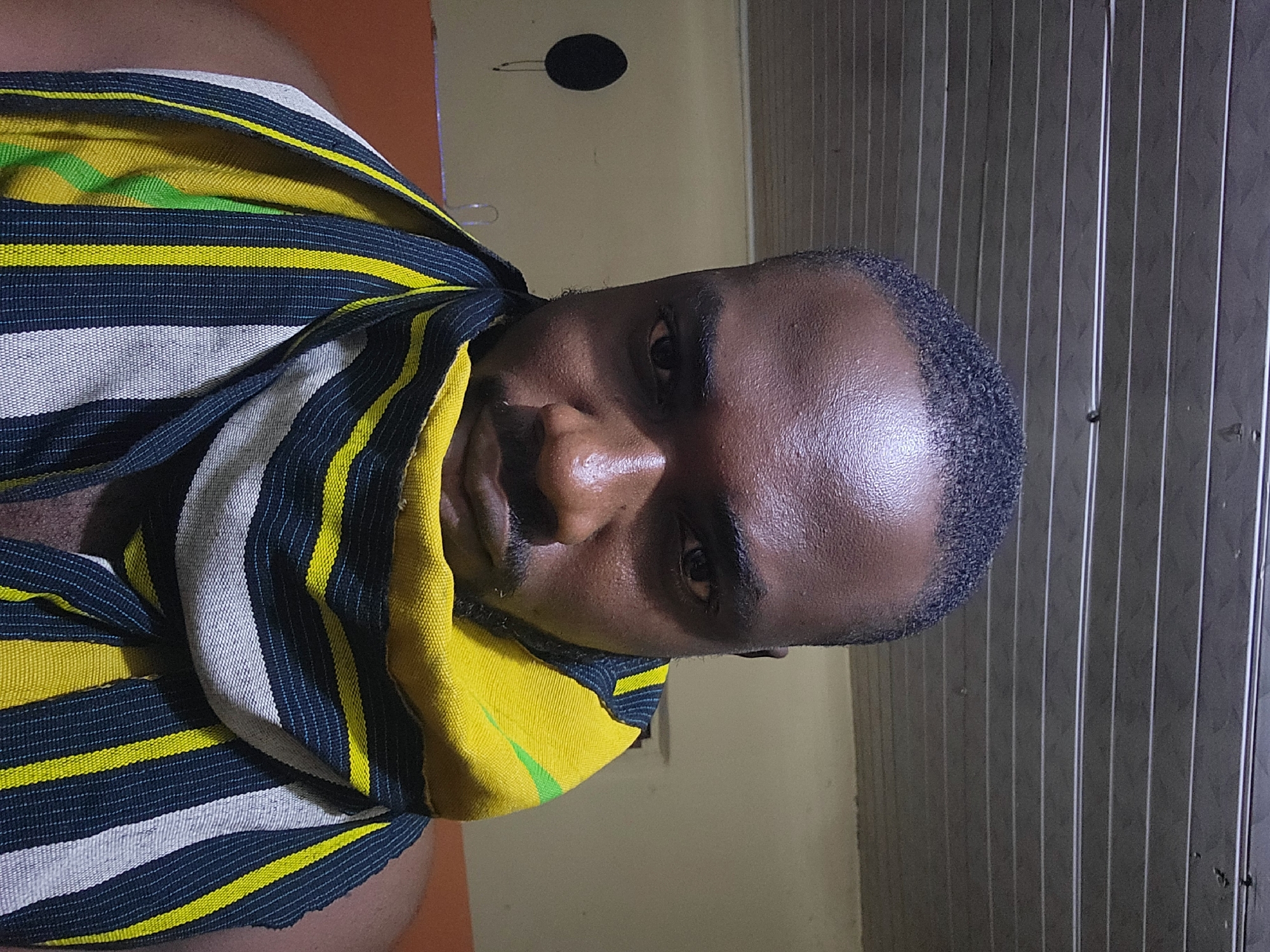 A close-up, head-and-shoulders portrait of a dark-skinned male individual is depicted indoors in the city of Jos, Nigeria. The man, approximately in his 20s or 30s, has short, dark, textured hair, a trimmed mustache, and a short beard. He looks directly into the camera with a neutral expression. A woven textile garment, possibly a scarf or traditional cloth, is draped around his neck and shoulders, featuring prominent stripes in yellow, dark blue, white, and green. His bare left shoulder is visible in the lower left quadrant. The background includes a ribbed, light-colored ceiling with horizontal panels. A light yellow wall is visible behind his left side, on which a dark, circular object hangs, with a thin dark line extending downwards from it. The wall behind his right side appears to be a slightly darker, possibly orange-brown hue. The scene is illuminated by an overhead light source, casting highlights on the man's forehead and nose.