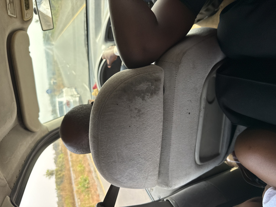 An interior view from a vehicle in Mayo, Nigeria, shows the back of a bald male driver's head and a gray fabric driver's seat, exhibiting multiple dark stains and wear. The driver's right hand grips the steering wheel, a light-colored watch on his wrist. To the right of the driver, the dark-skinned legs of a front passenger, clad in dark attire, are visible. To the left, the light-clad legs of another rear passenger are partially visible. Through the windshield, a two-lane asphalt road with a yellow median stripe extends into a hazy distance. A white sedan proceeds ahead and to the left. A blue circular road sign is visible further along the road. To the left, a railway track runs parallel to the road. Roadside vegetation appears dry and sparse under a bright, overcast sky.
