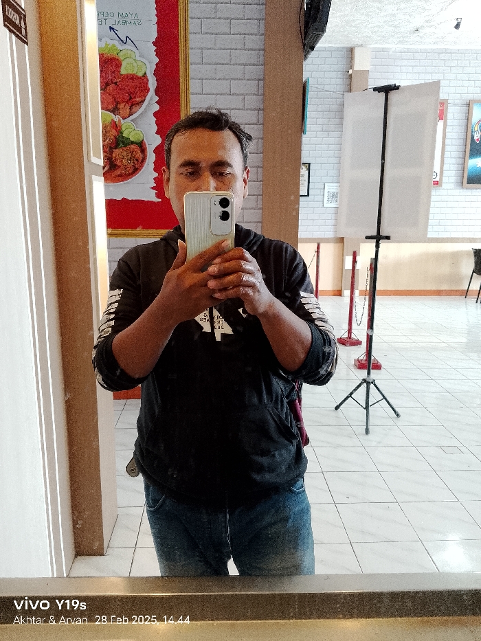 The image shows a man taking a selfie in front of a mirror in a restaurant in Patrol, Indonesia. He is wearing a black jacket and blue jeans and is holding a white smartphone in his right hand. His left hand is holding the phone steady. The phone is in portrait mode and the man is looking into the camera. The restaurant is decorated with white brick walls and the floor is tiled. There is a black stand with a white poster on it and a red and white barrier in the background. The image is taken from a slightly high angle and is blurry. The image is labeled with "vivo Y19s" and "Akhtar & Arvan 28 Feb 2025 14.44."