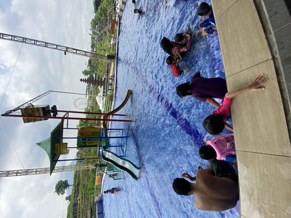 An outdoor swimming pool, possibly in a resort, is filled with people. A group of children and adults are clustered near the pool's edge, engaging in water play. A playground structure featuring a slide and climbing elements stands in the water at the other end. The backdrop showcases lush greenery and a distant Ferris wheel, hinting at an amusement park. The sky above is overcast.