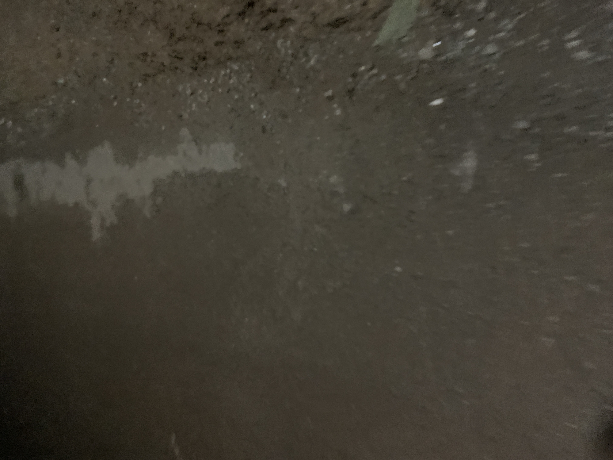 The image shows a close-up, low-angle view of a wet, dark, and textured surface, likely a street or path, at night. The surface appears to be a mixture of asphalt or concrete with small stones and debris. There is a reflective patch of water, possibly a puddle, running diagonally across the upper-middle portion of the frame, creating a lighter, elongated shape against the darker surroundings. The lighting is dim, with some bright spots that could be reflections from streetlights or other light sources. The overall impression is one of a damp, possibly neglected, urban environment.