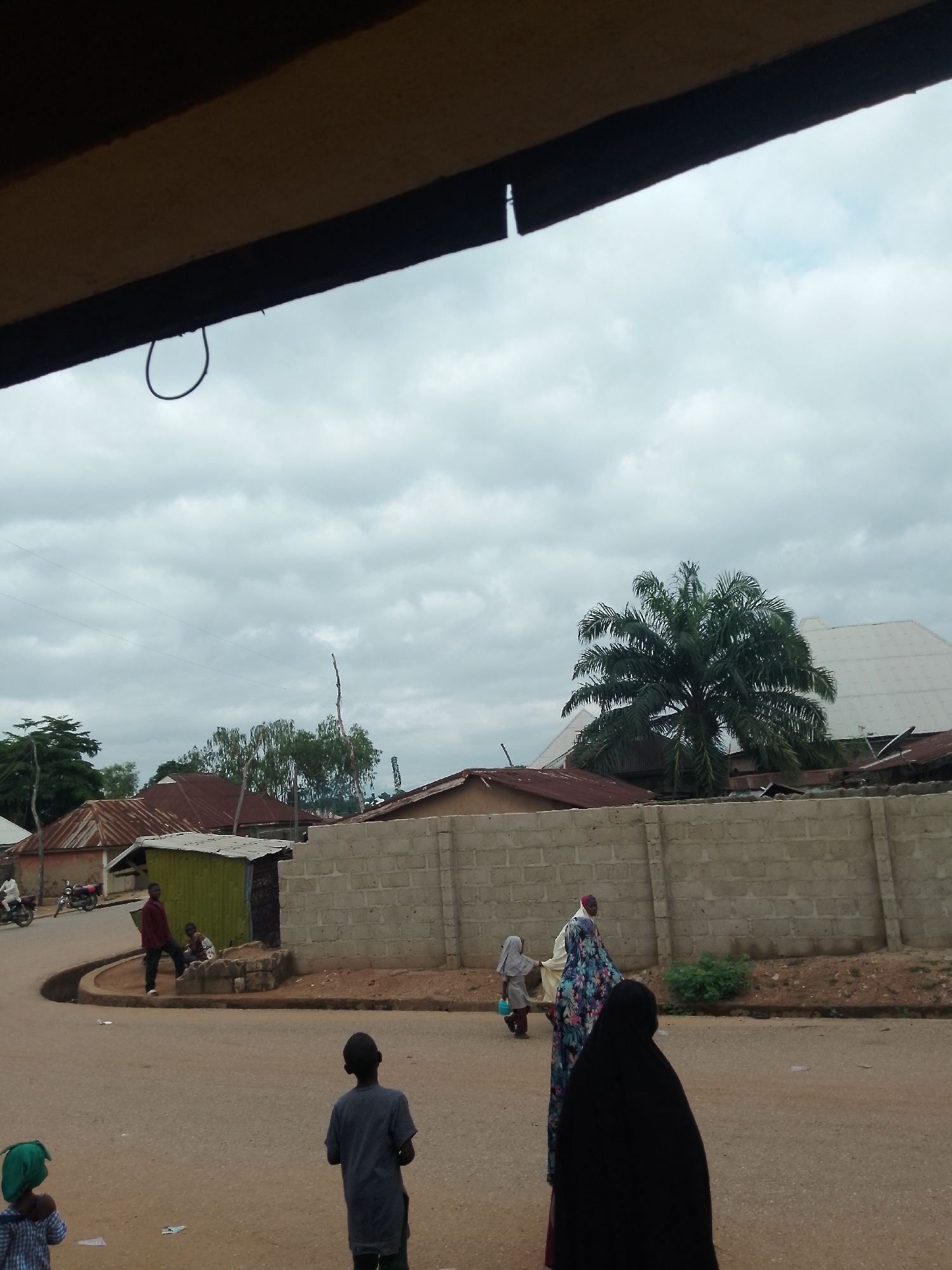 A medium shot captures a cloudy, overcast day in a village setting, with a view of a street, some buildings, and people. In the foreground, a young person wearing a blue and white checkered shirt and a green headscarf is facing away from the camera. To their right, another young person in a dark grey shirt is also facing away.
Further down the dusty road, a woman in a colorful patterned dress and headscarf is walking away from the camera, followed by another person in a black hijab. To the left, a man in a maroon shirt stands near a small green corrugated metal structure. Two children are seated near a stone structure behind the man.
In the background, a concrete block wall runs horizontally across the frame. Beyond the wall, several buildings with rusty metal roofs are visible, along with a large palm tree. A white roof of a building is also apparent. The street is mostly bare, with a few scattered pieces of litter. A motorcycle is parked on the left side of the road.