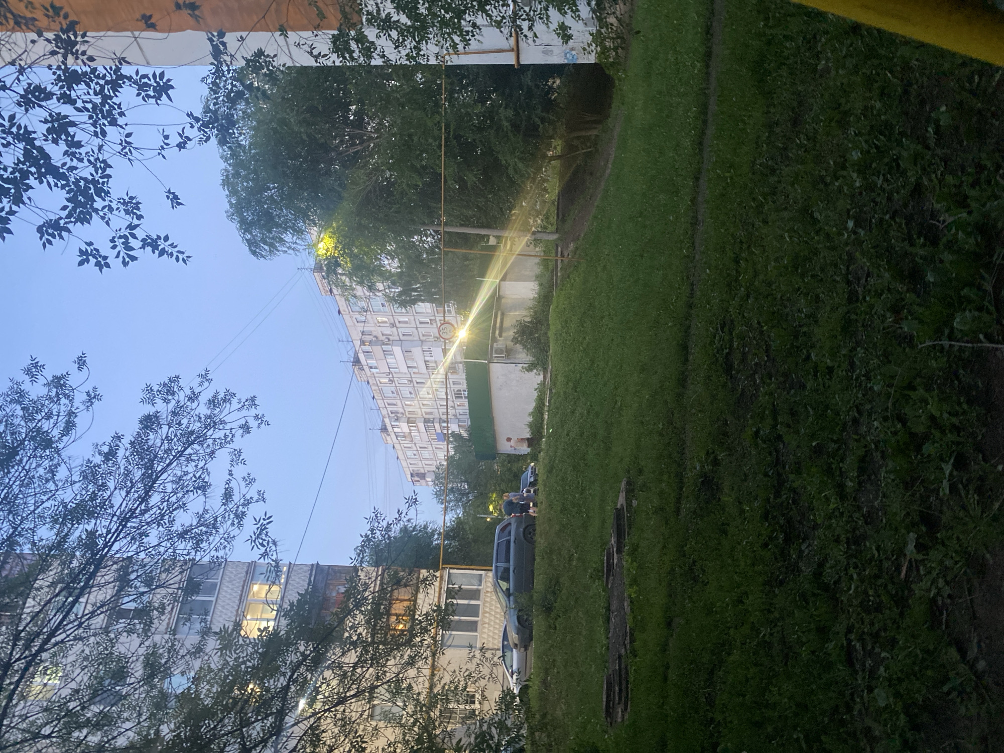 The image depicts a residential area in Samara, Russia, during twilight. In the foreground, a grassy embankment slopes downward, with a patch of dark soil and greenery at the very bottom. Several concrete manhole covers are visible on the embankment. On the left, a tall, multi-story apartment building with visible windows, some illuminated, is partially obscured by tree branches. A few cars are parked near the building. In the middle ground, another, longer apartment building stretches across the frame, with many windows. A bright light source, possibly a street lamp, shines from behind a tree in front of this building, creating a strong lens flare and visible light rays. Several people are standing near a car in front of the longer apartment building. A large tree with dense foliage stands between the apartment buildings. The sky is a clear, pale blue.
