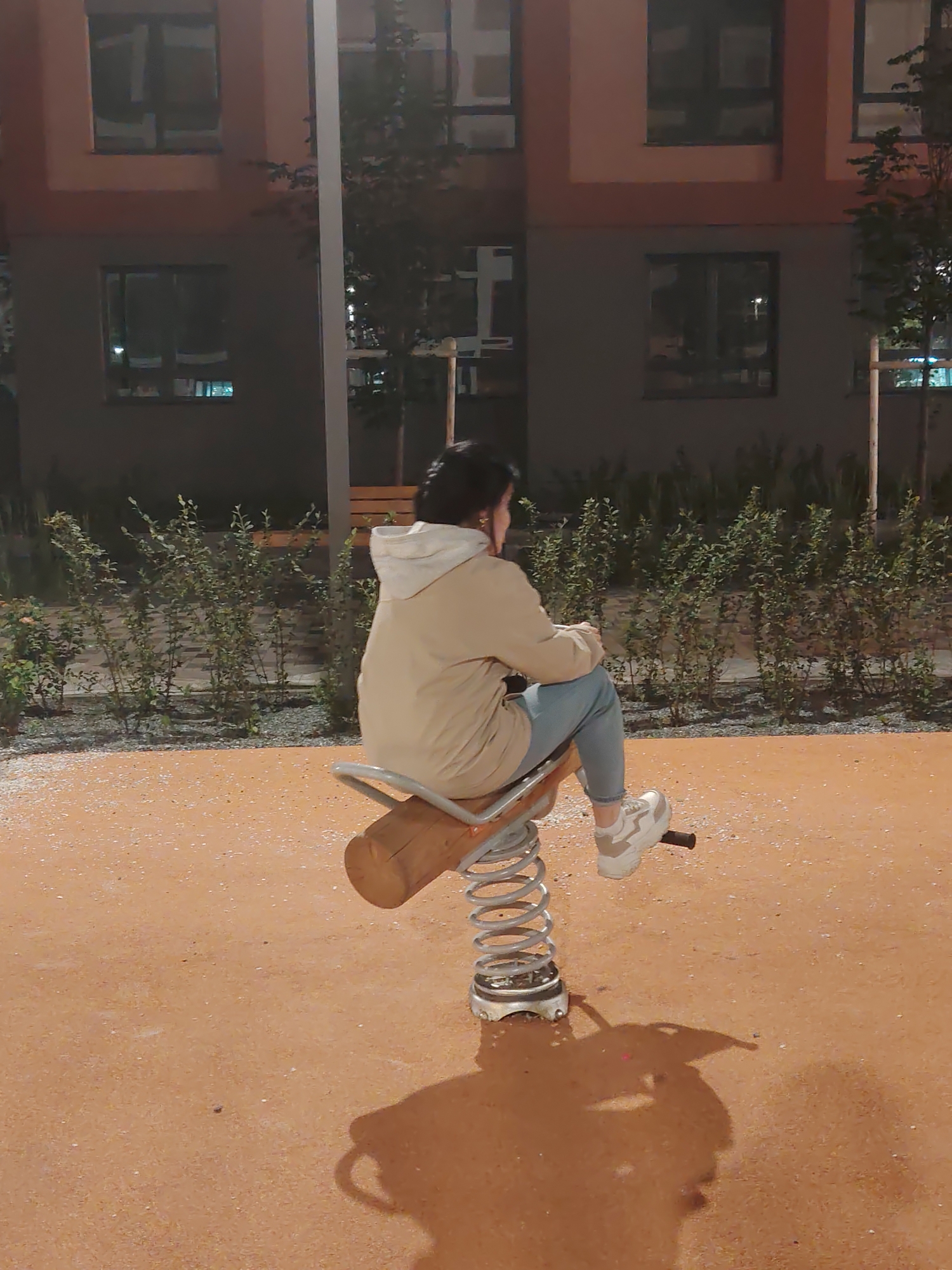 In Yekaterinburg, Russia, a young woman is captured from behind, sitting on a wooden spring rider in a park at dusk. She is wearing a light-colored hoodie and blue jeans, with white and beige sneakers. The rider itself is made of wood and metal, with a coiled spring. The ground beneath is covered in an orange, sandy material, speckled with white pebbles. In the background, a modern building with dark windows and balconies is visible, along with lush green bushes and trees. A street light casts a shadow of the woman and the rider onto the ground, creating an elongated silhouette. The overall scene depicts a quiet moment in an urban park during the evening hours.
