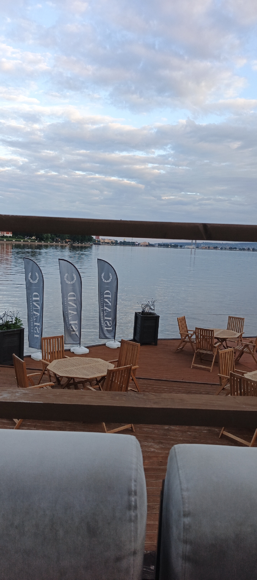 The image shows an outdoor seating area with wooden tables and chairs on a wooden deck, adjacent to a body of water under a cloudy sky. Three vertical flags with a logo are prominently displayed. In the foreground, the backs of two light-colored upholstered seating units are visible. The setting appears to be a waterfront establishment, possibly a restaurant or lounge. The scene is calm and empty of people. The location is identified as Votkinsk, Russia.