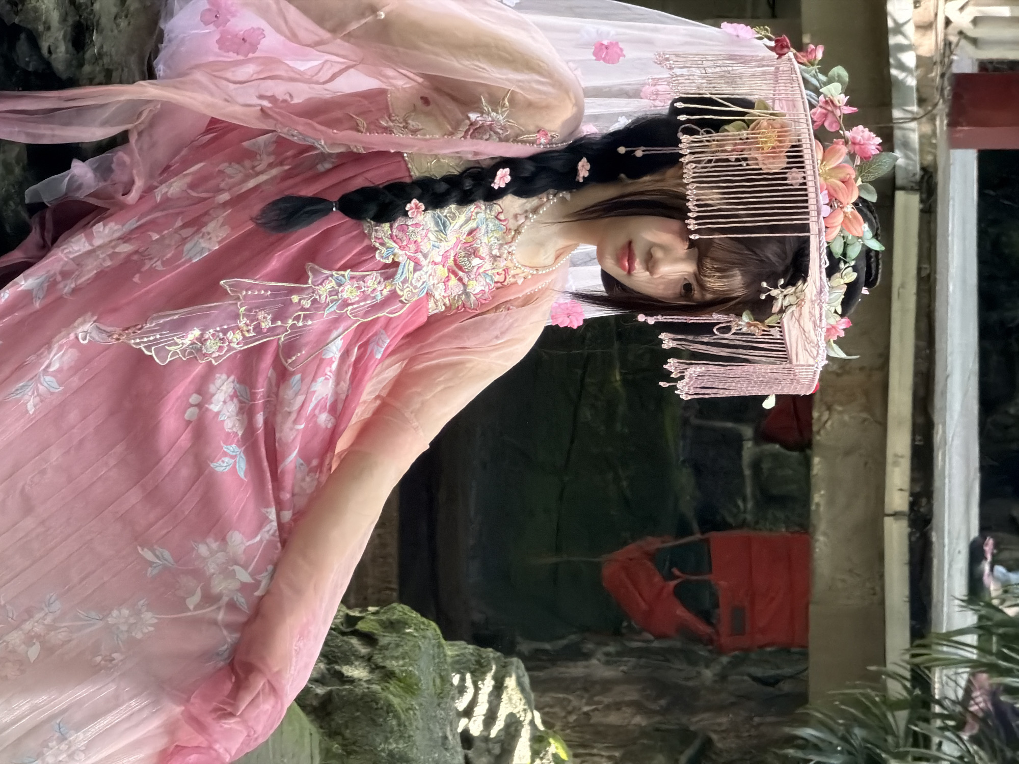 A young woman adorned in a pink traditional Chinese Hanfu dress with floral embroidery and pearl necklaces is posing outdoors. She wears an elaborate headdress featuring flowers and tassels, and her hair is styled in a braid with flower accents. The background suggests a garden or courtyard setting with stone structures and foliage, possibly a traditional Chinese garden in Yuzhong District, China. The lighting indicates it is daytime, and the overall mood is serene and artistic.